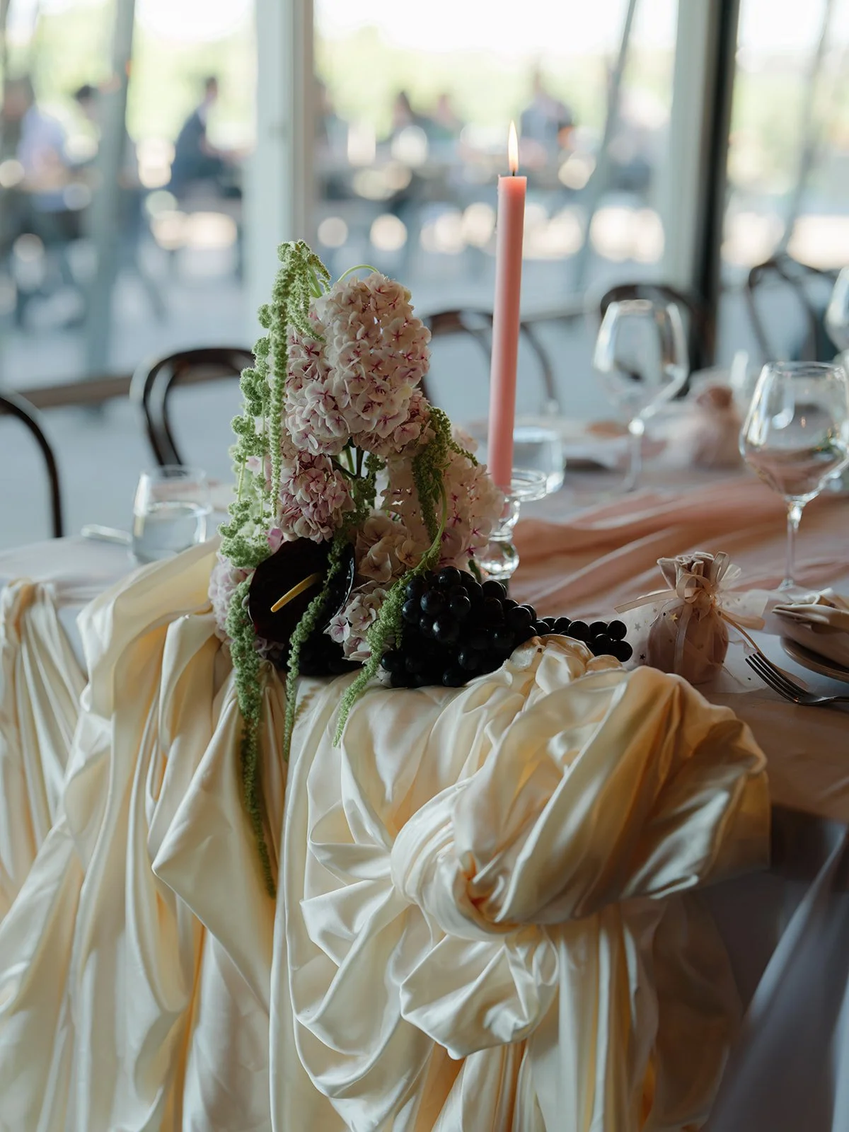 Table decoration with flowers, a pink candle, grapes, and a small wrapped gift at a formal event.