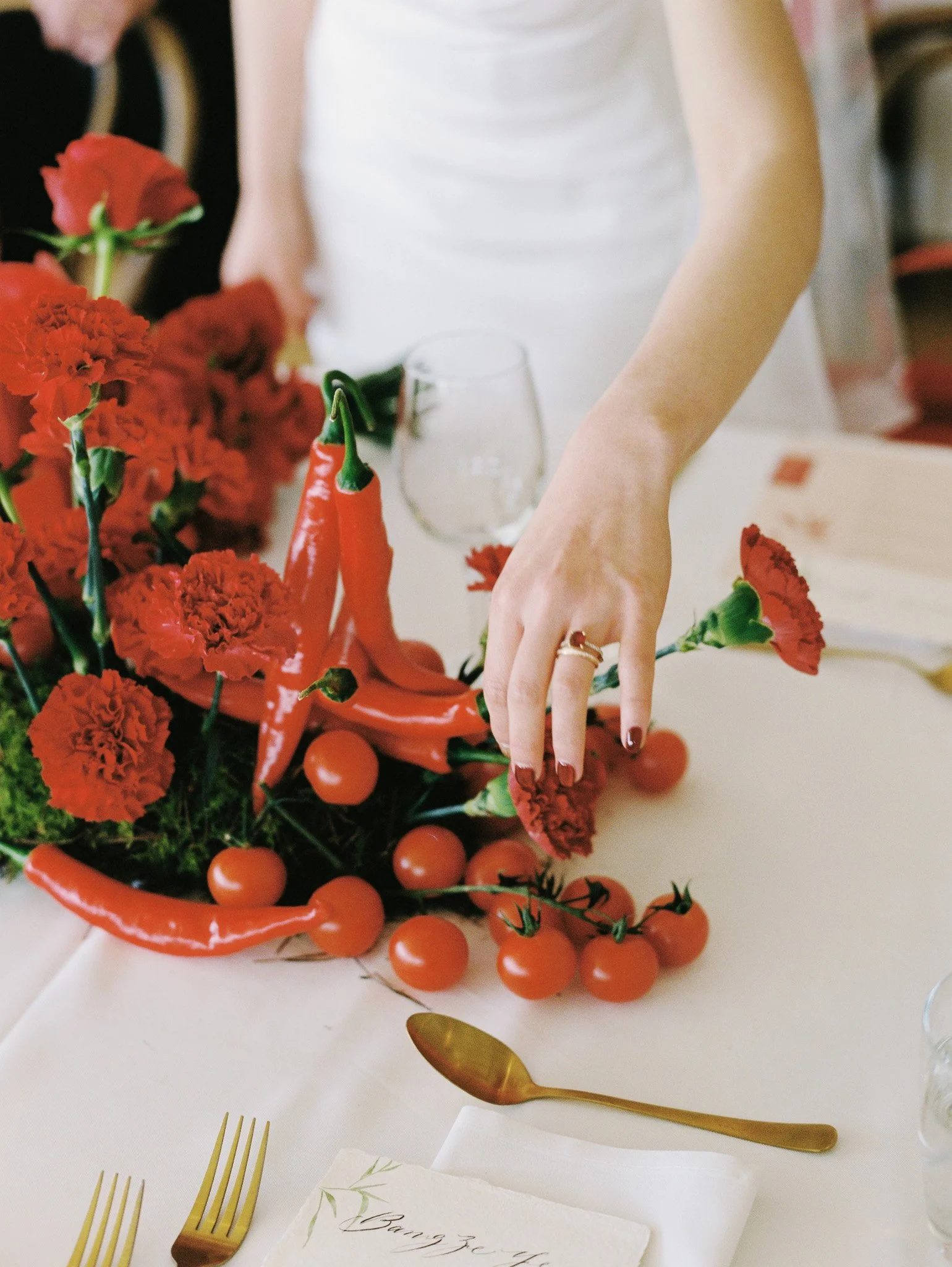 A table setting with gold utensils, a menu card, and a floral arrangement made of red carnations, red chili peppers, and cherry tomatoes.