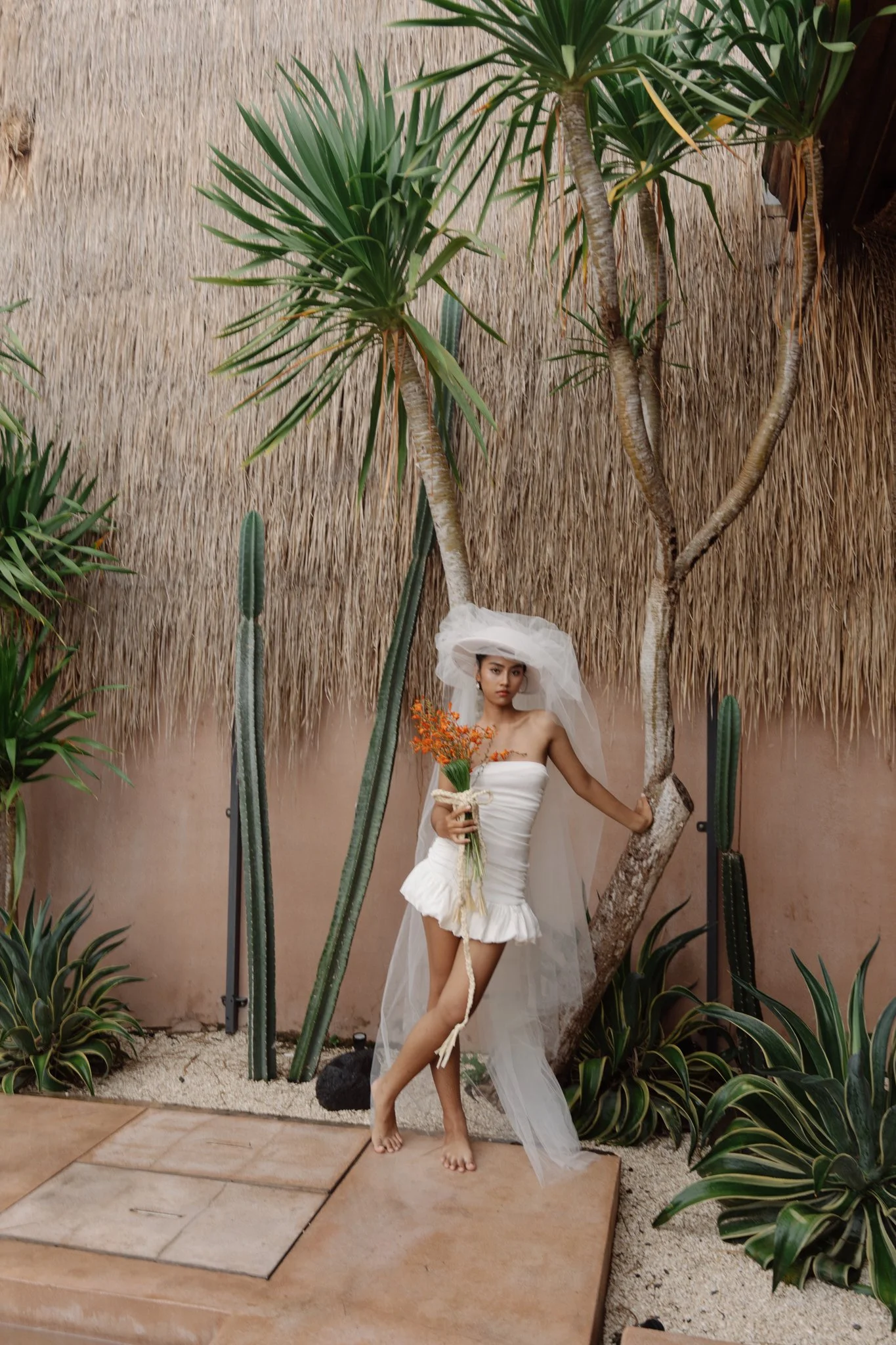A woman in a white dress and veil holding a bouquet of orange flowers, standing among tall desert plants and cacti against a thatched wall background.