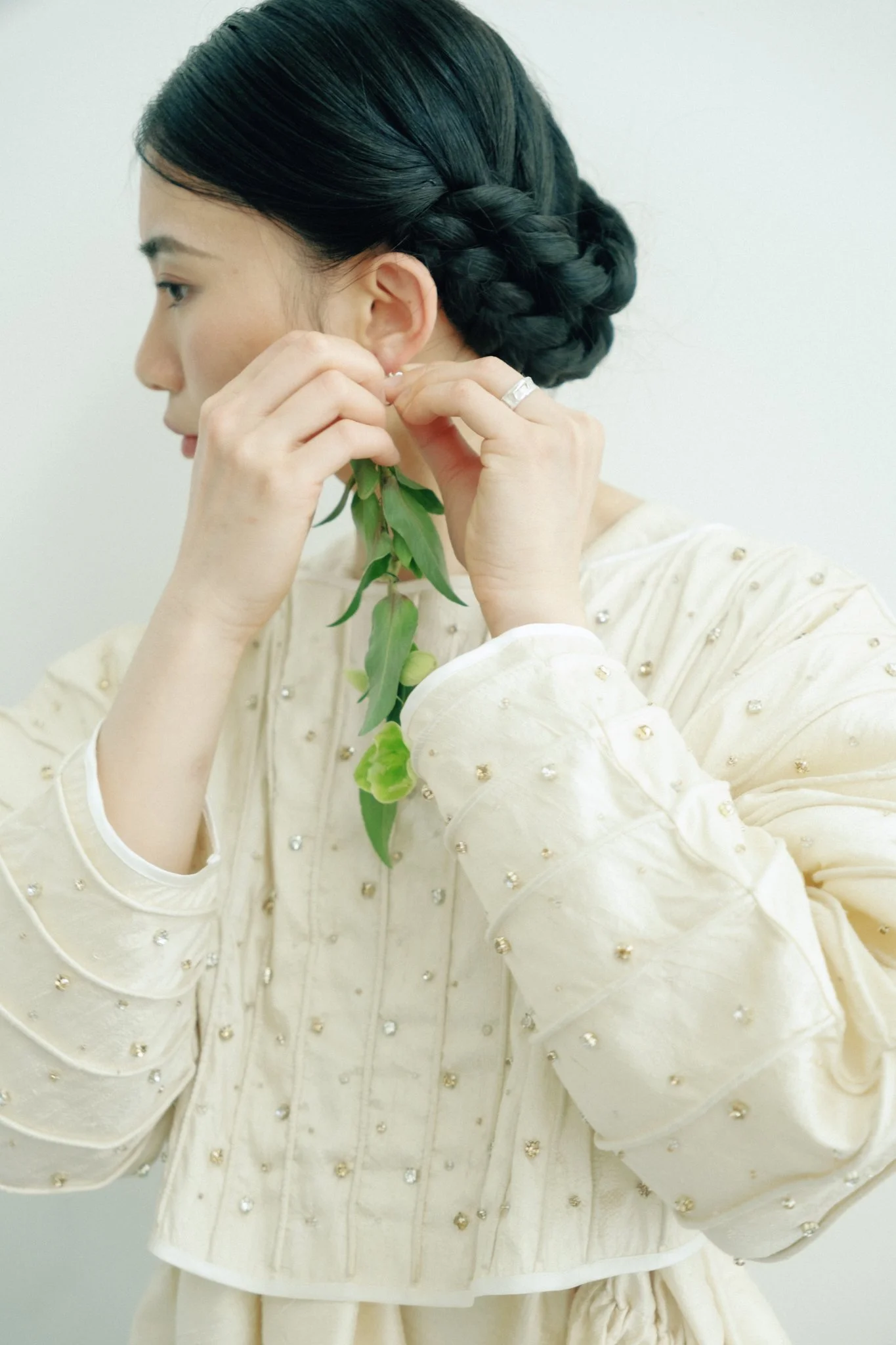 A woman with dark hair styled in a braided bun, wearing a cream-colored top with pearl-like embellishments, is putting on an earring and holding a branch with green leaves.