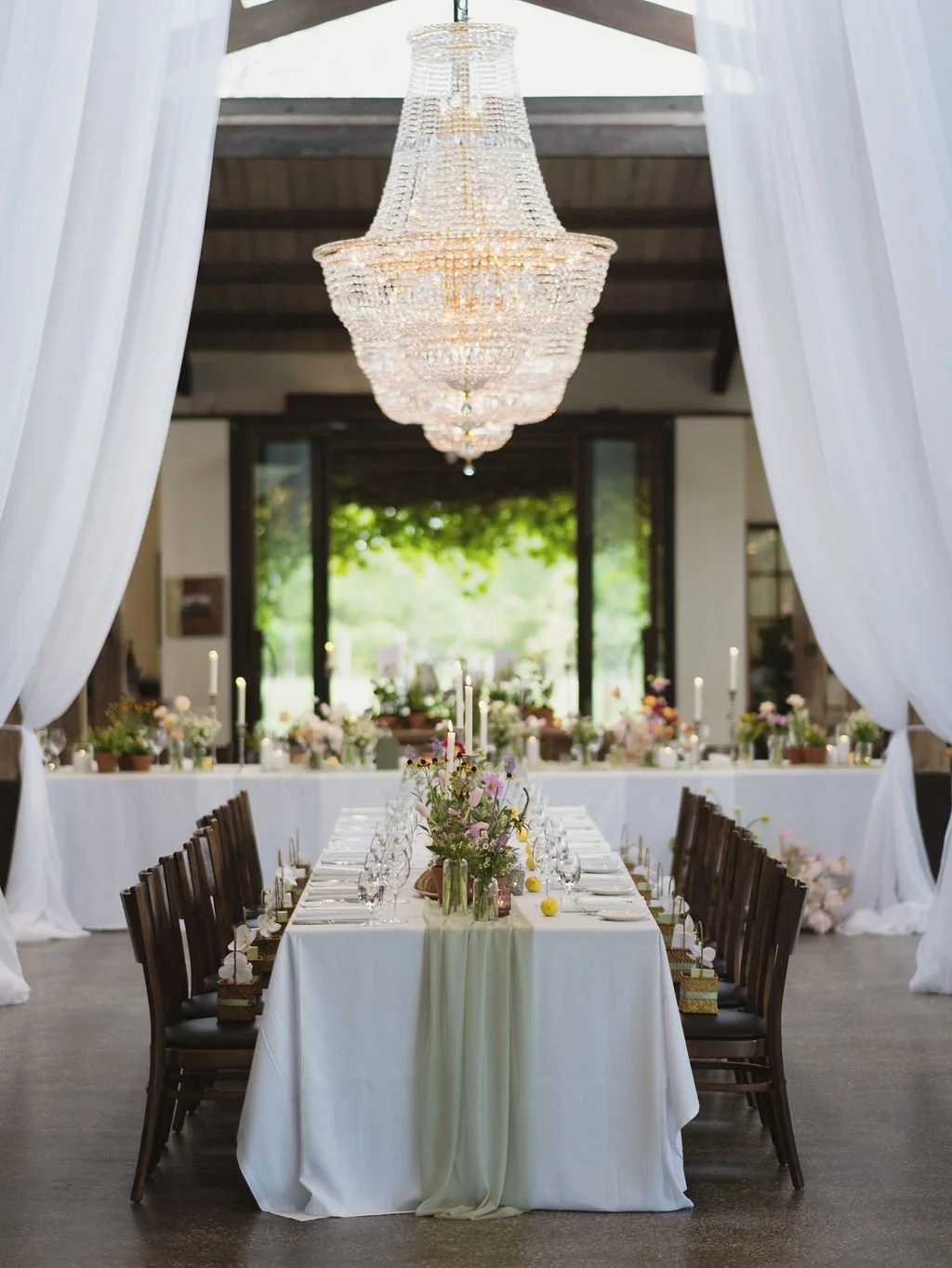 Elegant wedding reception setting with a chandelier, long tables with white tablecloths, floral centerpieces, candles, and surrounded by dark wooden chairs. White curtains frame the scene, with a view of greenery outside.