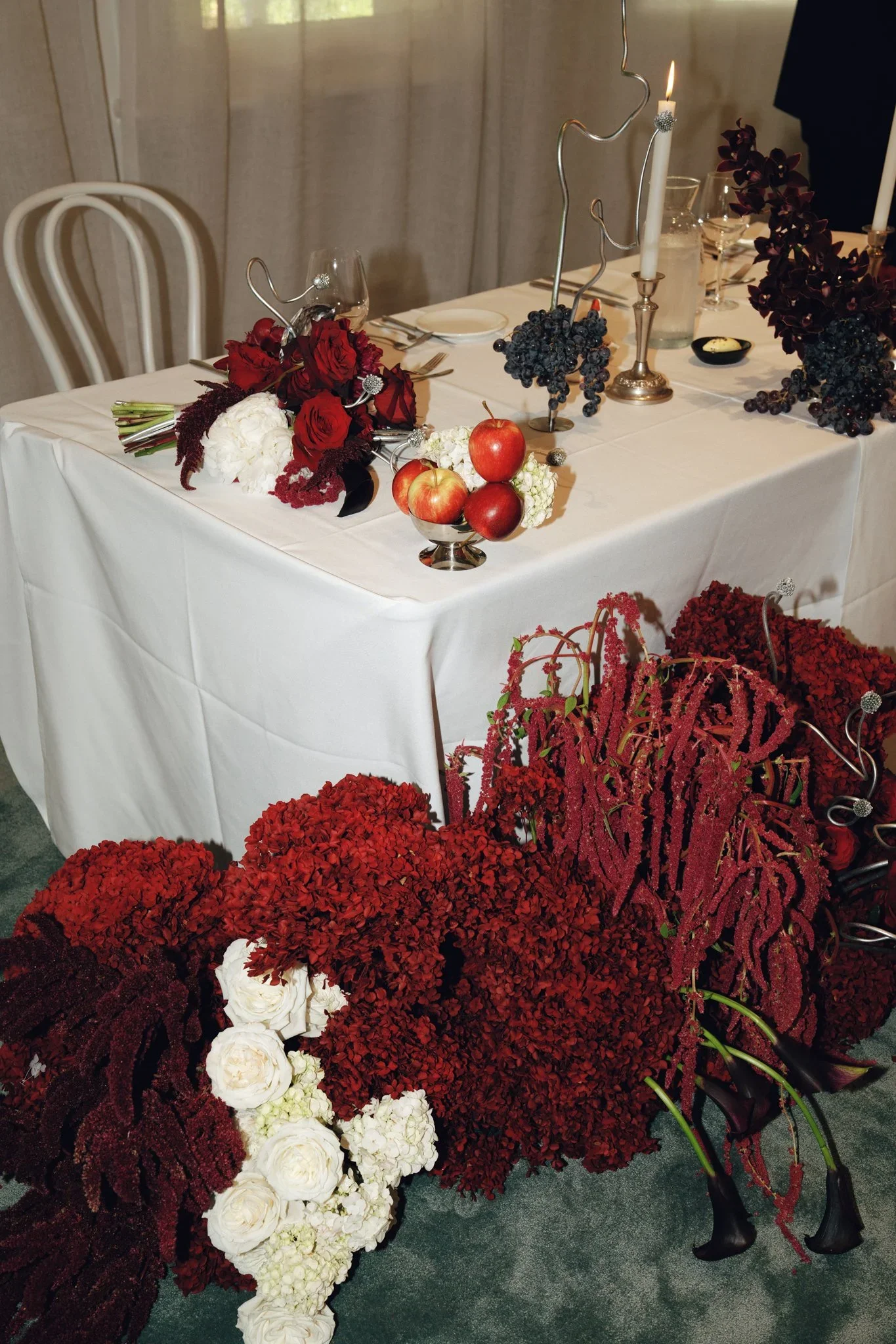 A decorated dining table with red, white, and dark purple flowers, bunches of grapes, red apples, candlesticks with lit candles, glassware, and tableware. The table is covered with a white tablecloth and has chairs nearby.