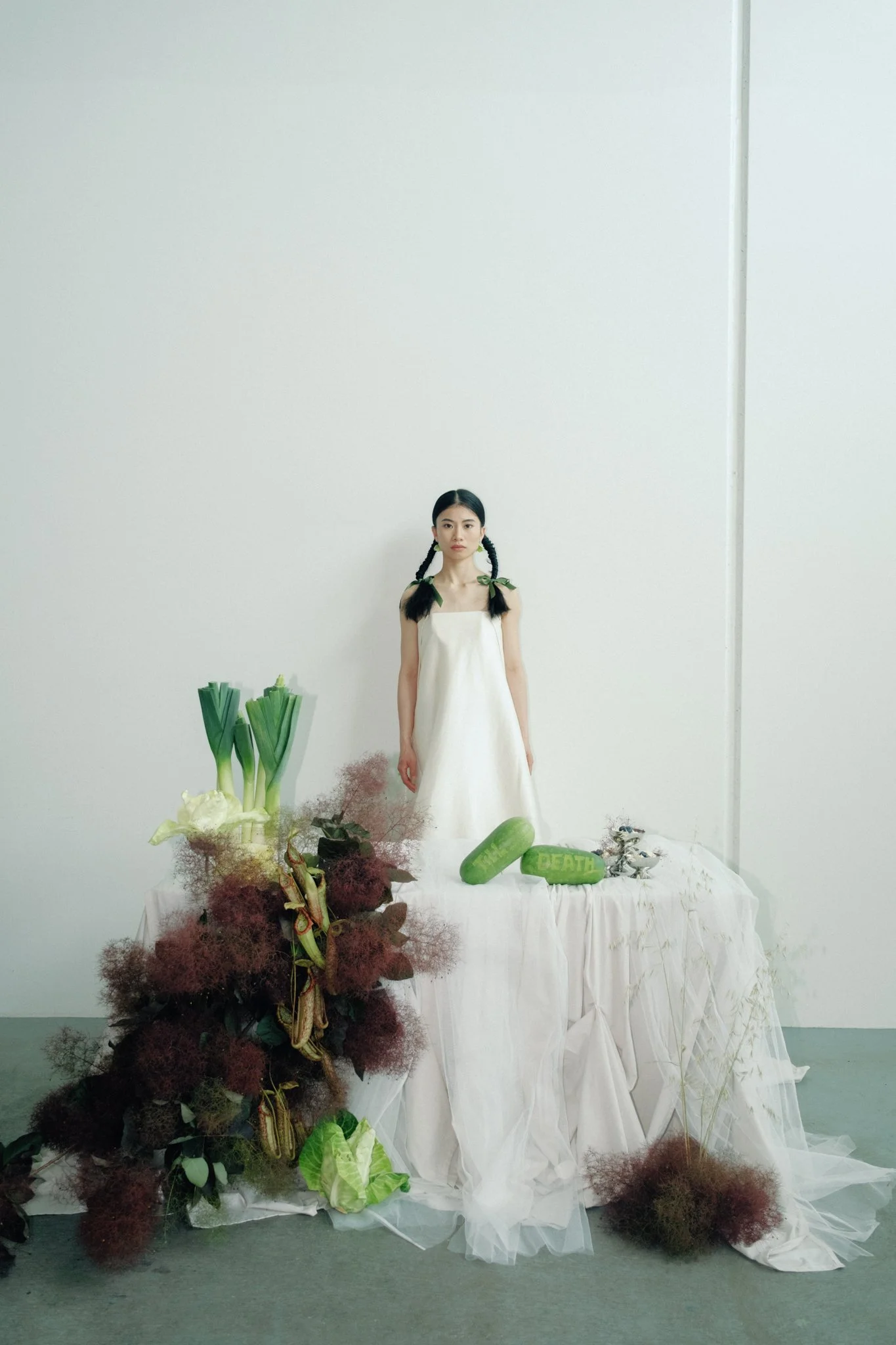 A woman in a white dress standing behind a table covered with a white cloth, surrounded by various vegetables and plants, including leeks, cucumbers, and leafy greens, in an indoor setting.
