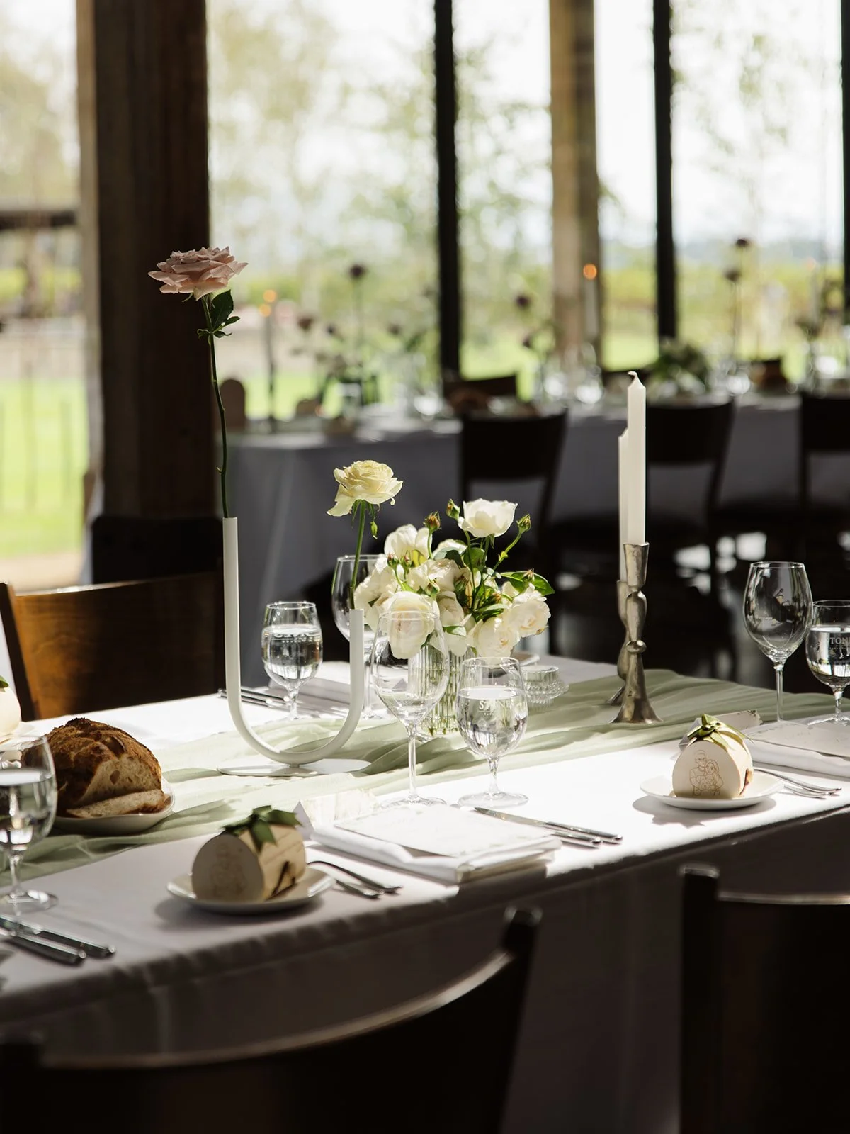 Elegant dining table set with white flowers, candles, wine glasses, bread, and a small ornament, in a well-lit room with large windows and natural light.
