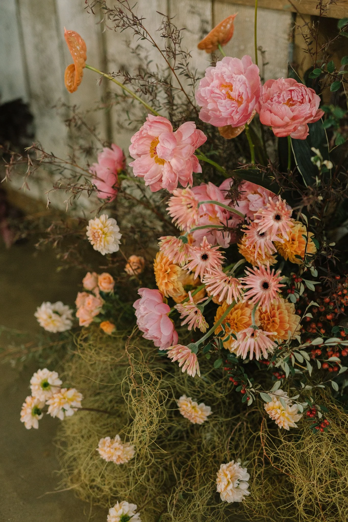A floral arrangement with pink peonies, orange dahlias, and smaller white and pink flowers, accented with dark reddish-brown stems and greenery, set against a rustic background.