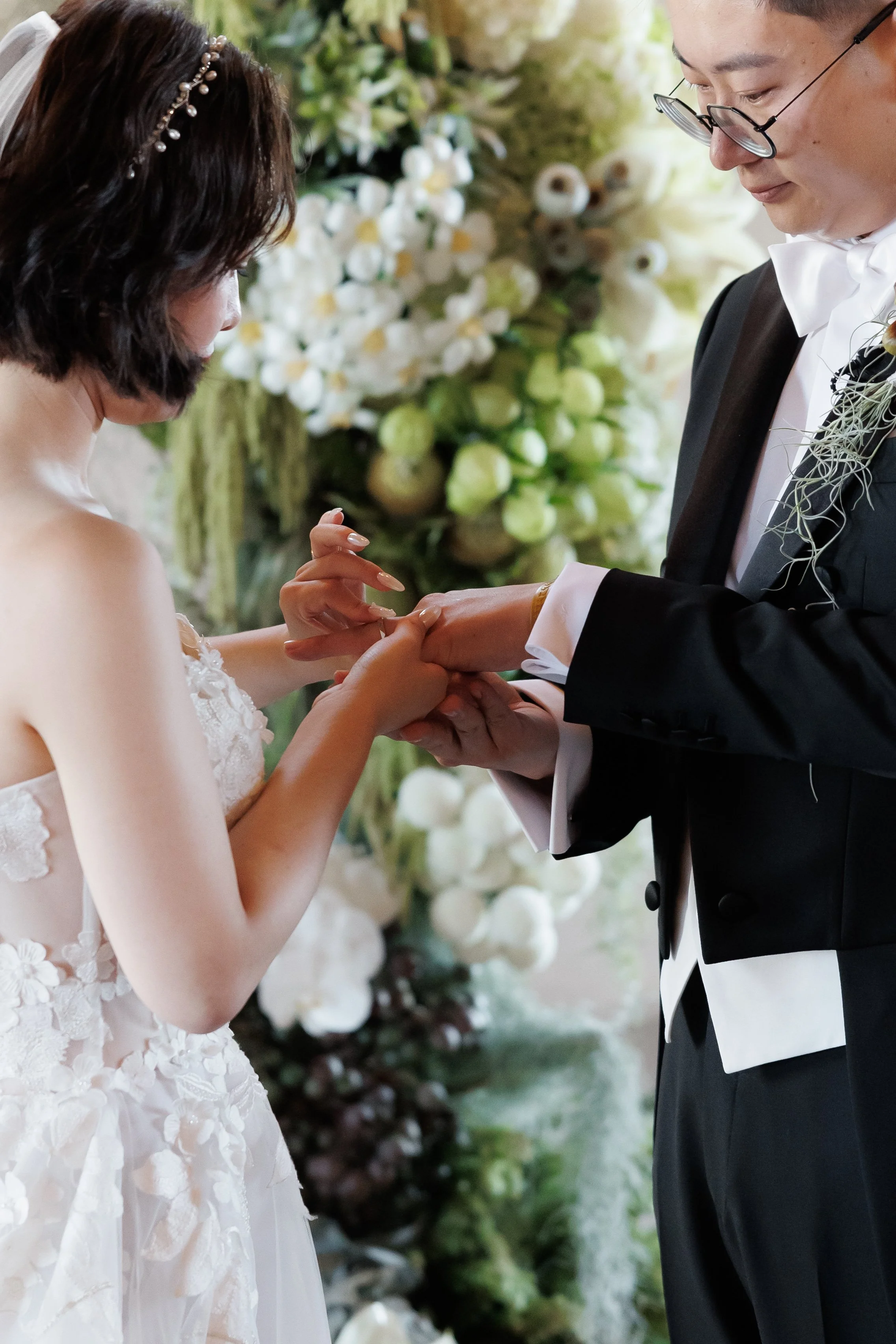 A bride and groom exchanging rings during their wedding ceremony, with white floral decorations in the background.