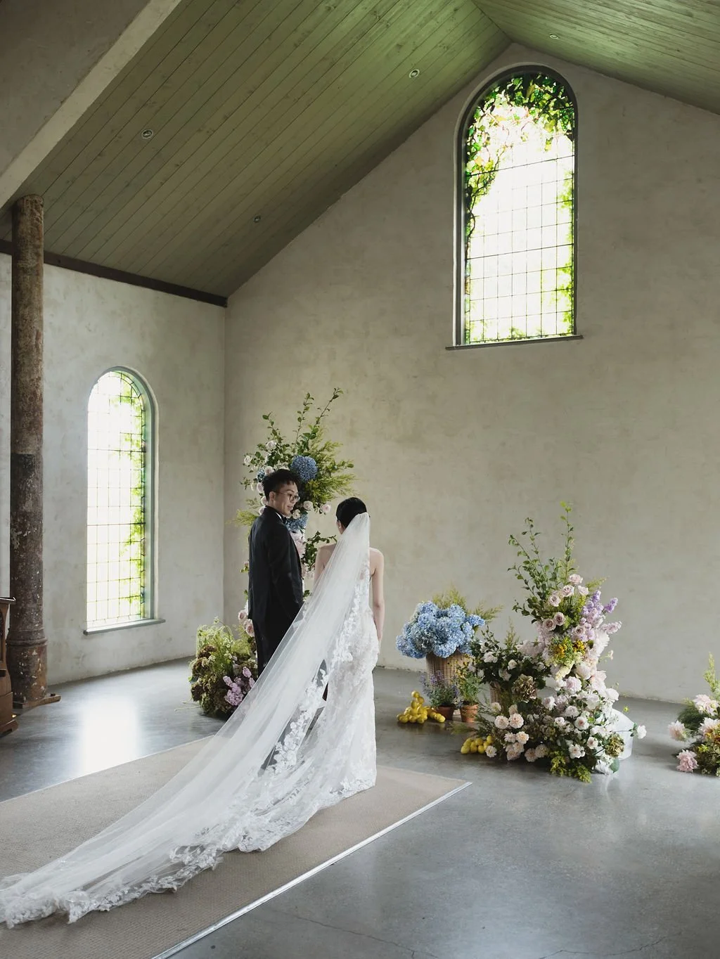 A bride and groom stand together during their wedding ceremony inside a minimalistic church with arched windows and floral arrangements.