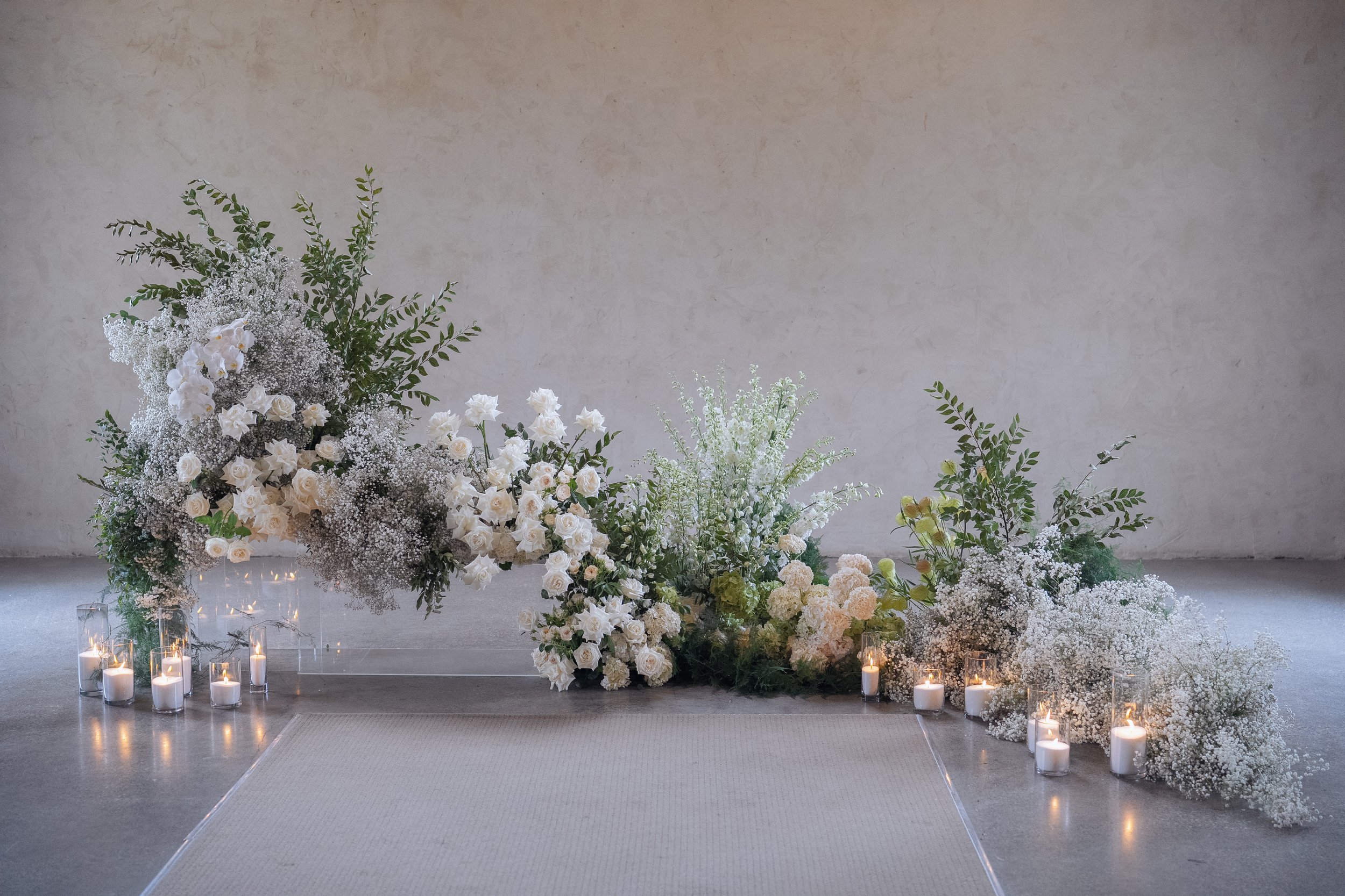 Elegant white flower arrangement with candles in glass holders on the floor, set against a plain wall, likely for a special event or wedding decor.