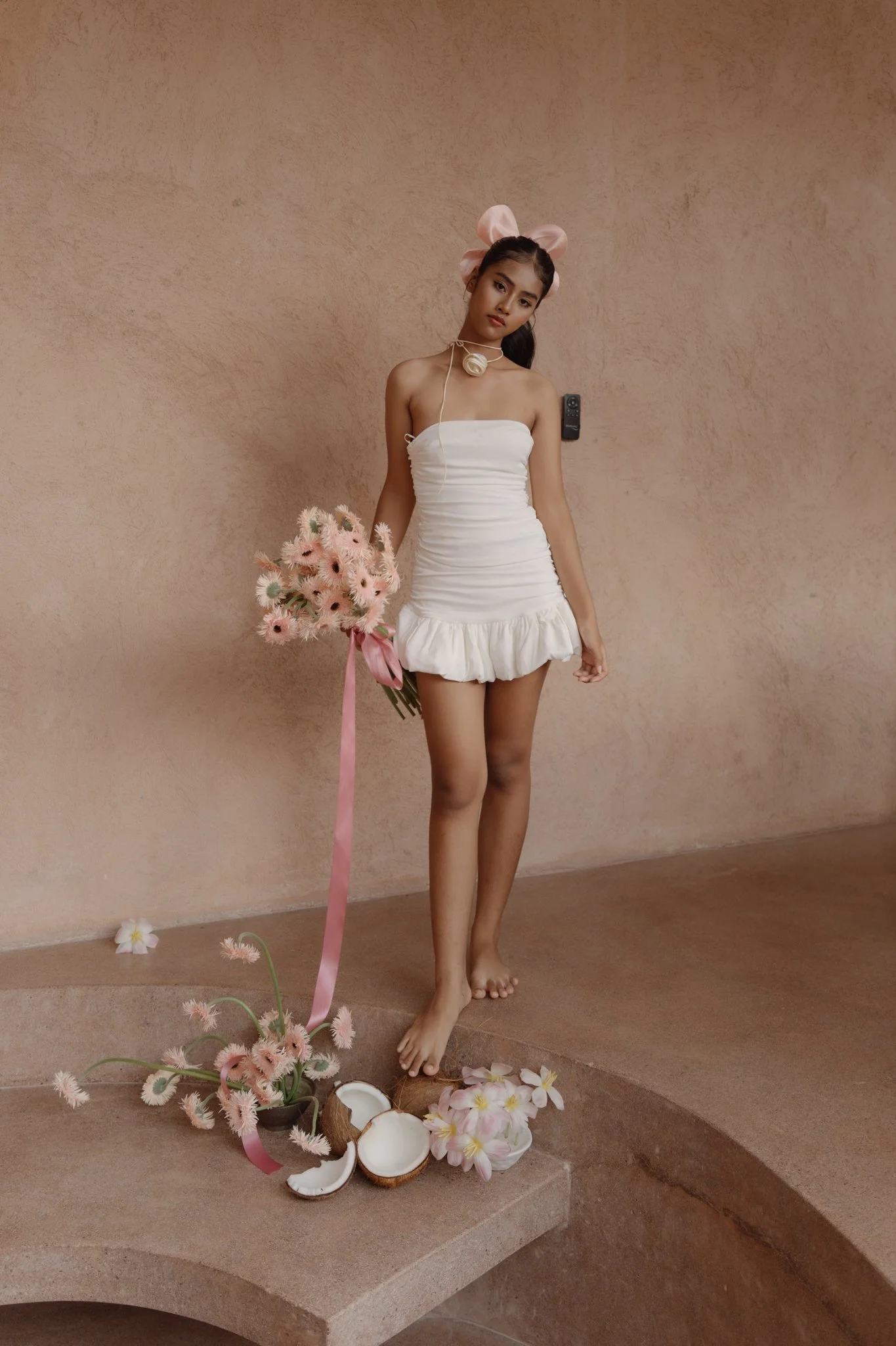 A young woman in a white dress holding a bouquet of pink flowers, standing barefoot on stairs against a peach-colored wall, surrounded by scattered flowers and broken coconut shells.