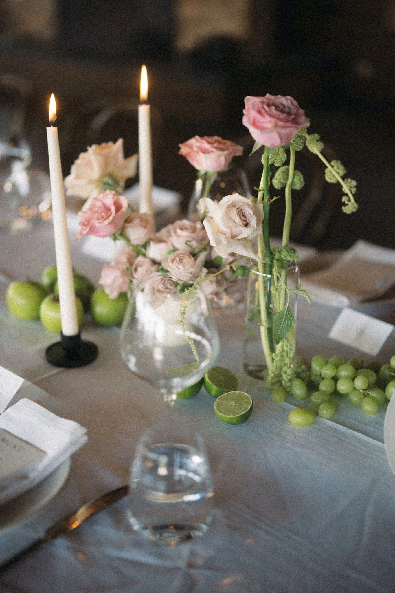 A table setting with pink and beige roses in glass vases, two lit white candles, green grapes, lime halves, and white napkins.