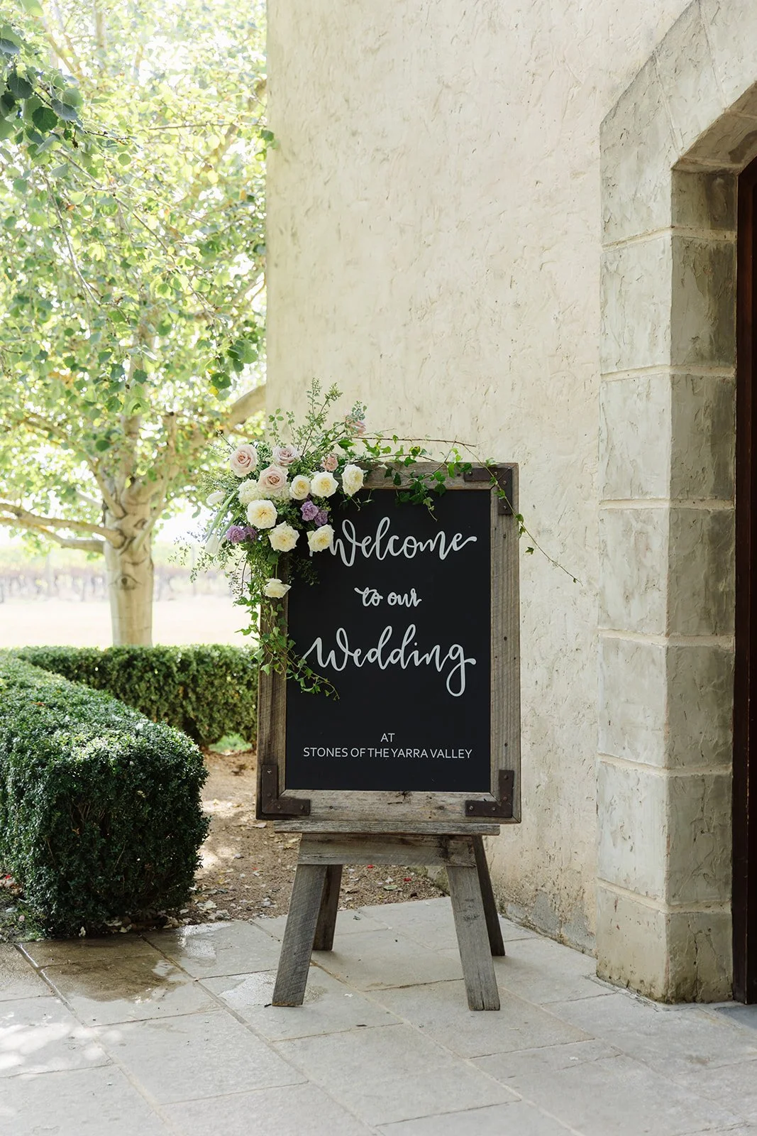A welcoming wedding sign decorated with flowers, positioned on stands outside a building with stone walls, with a tree and greenery in the background.