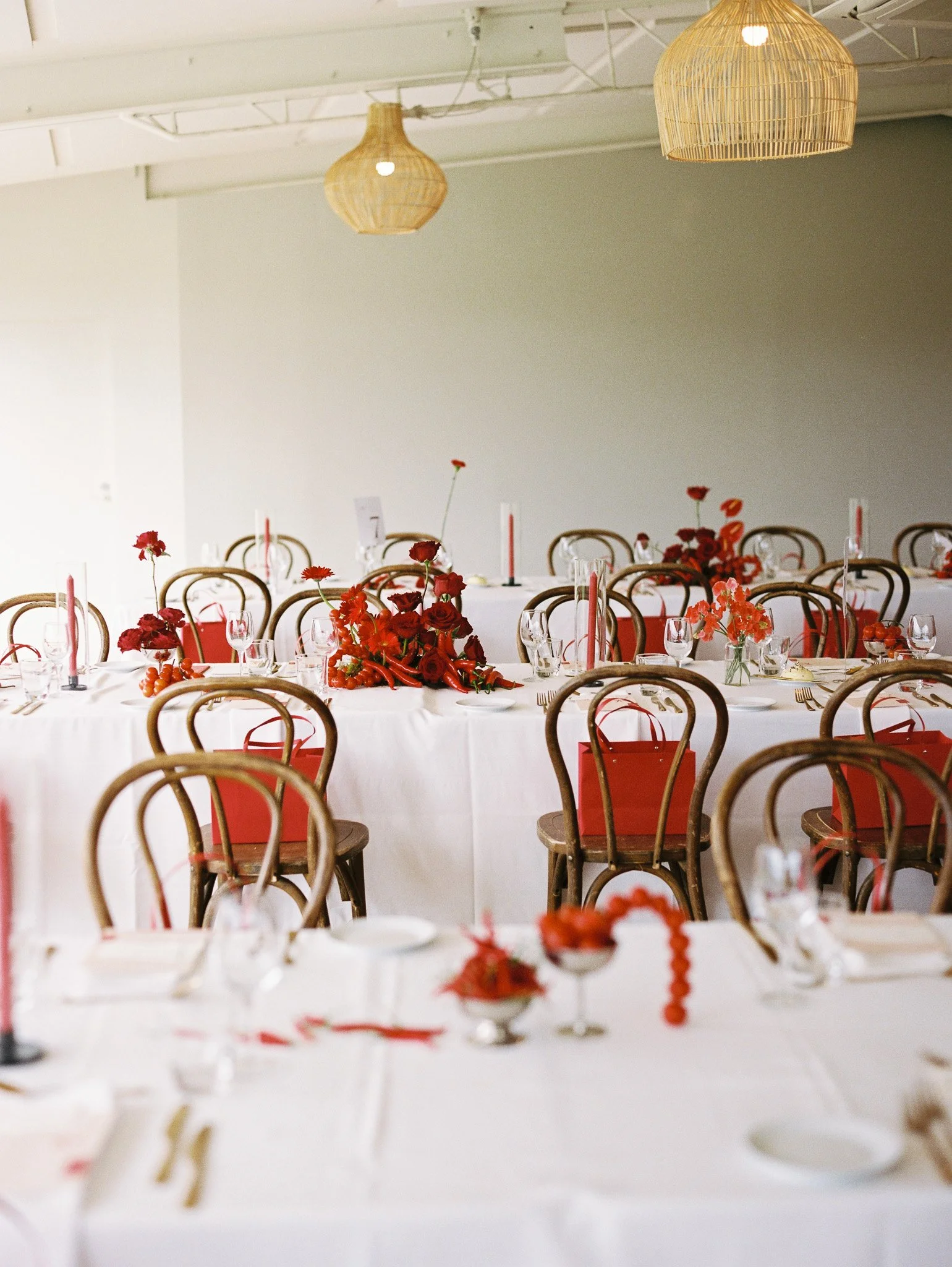 A decorated event table with red flowers, candles, wine glasses, and party favors, set under two hanging wicker lamps in a spacious hall with white walls.