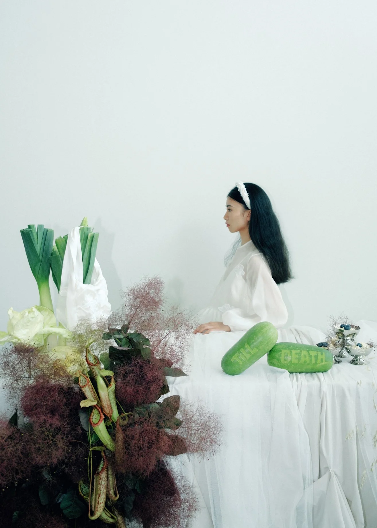 A woman with long black hair and a white headband sits in profile at a table decorated with green vegetables, flowers, and arts and crafts. Two watermelons with the words 'THE' and 'BEARTH' are on the table, along with a silver cup filled with bluebe