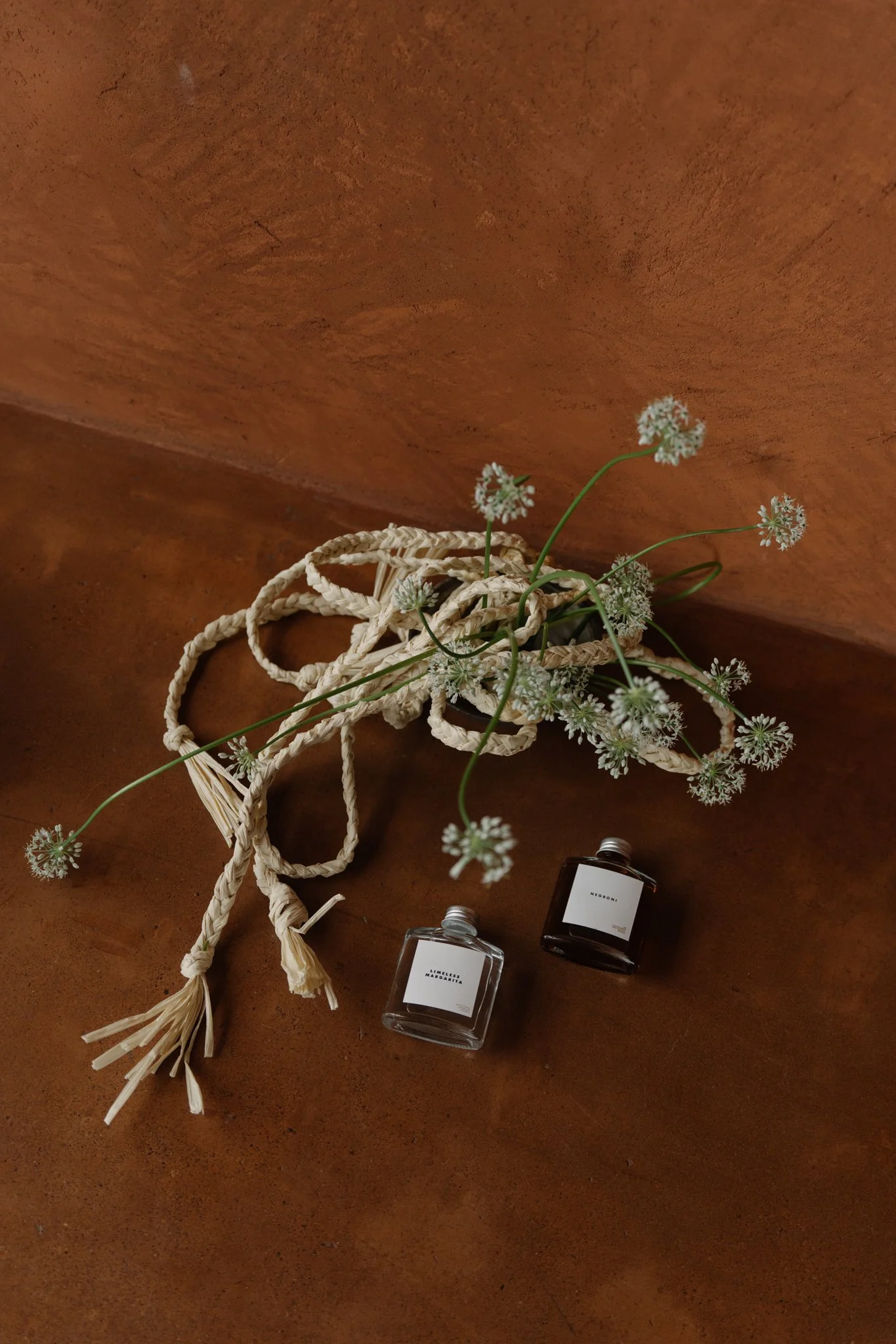 A small bouquet of white flowers with long green stems in a woven basket, two bottles of perfume labeled 'LIMELSS' and 'NEREONE,' and some white rope on a wooden surface.