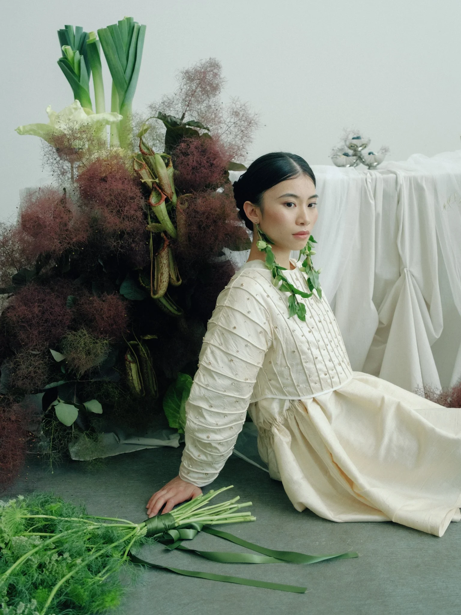 A woman dressed in a light-colored dress with green leaves around her neck, sitting on the floor beside a large arrangement of mixed green plants, flowers, and greenery on a gray surface against a plain white wall.