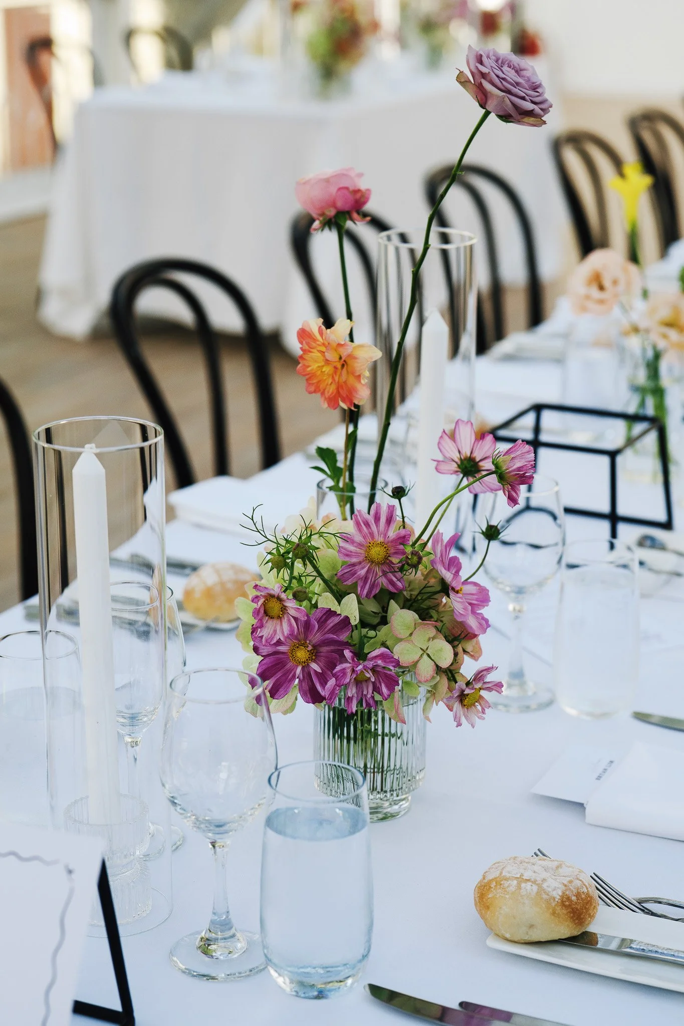A table decorated with a bouquet of pink, purple, and peach flowers in a glass vase, surrounded by clear glassware and candles, set for an event in a bright, well-lit room.