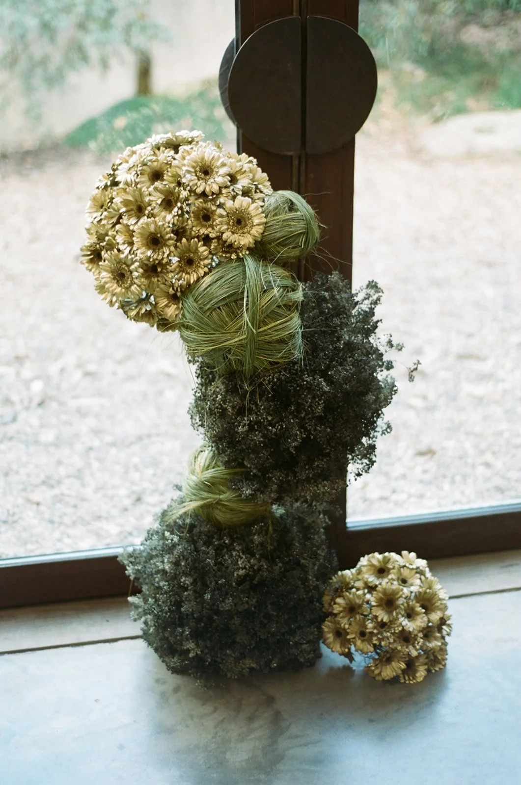 A decorative floral arrangement with dried flowers, including white daisies, and green and dark foliage, placed on a windowsill with a view outside.