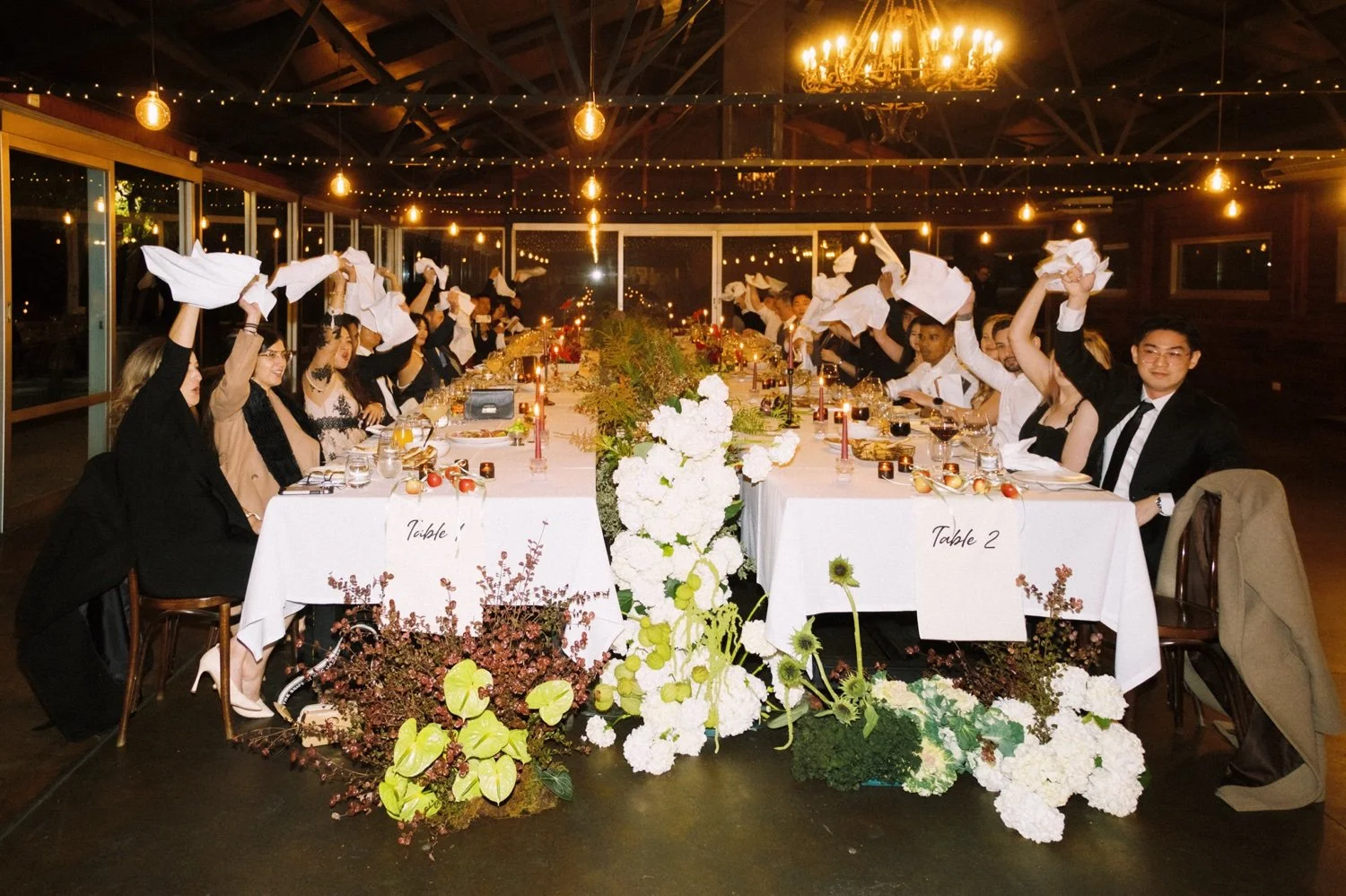 A formal dinner event in a decorated indoor venue with a long white table, floral centerpieces, candles, and string lights. People are seated on both sides, raising napkins in a celebratory gesture. The room has warm lighting and large windows.