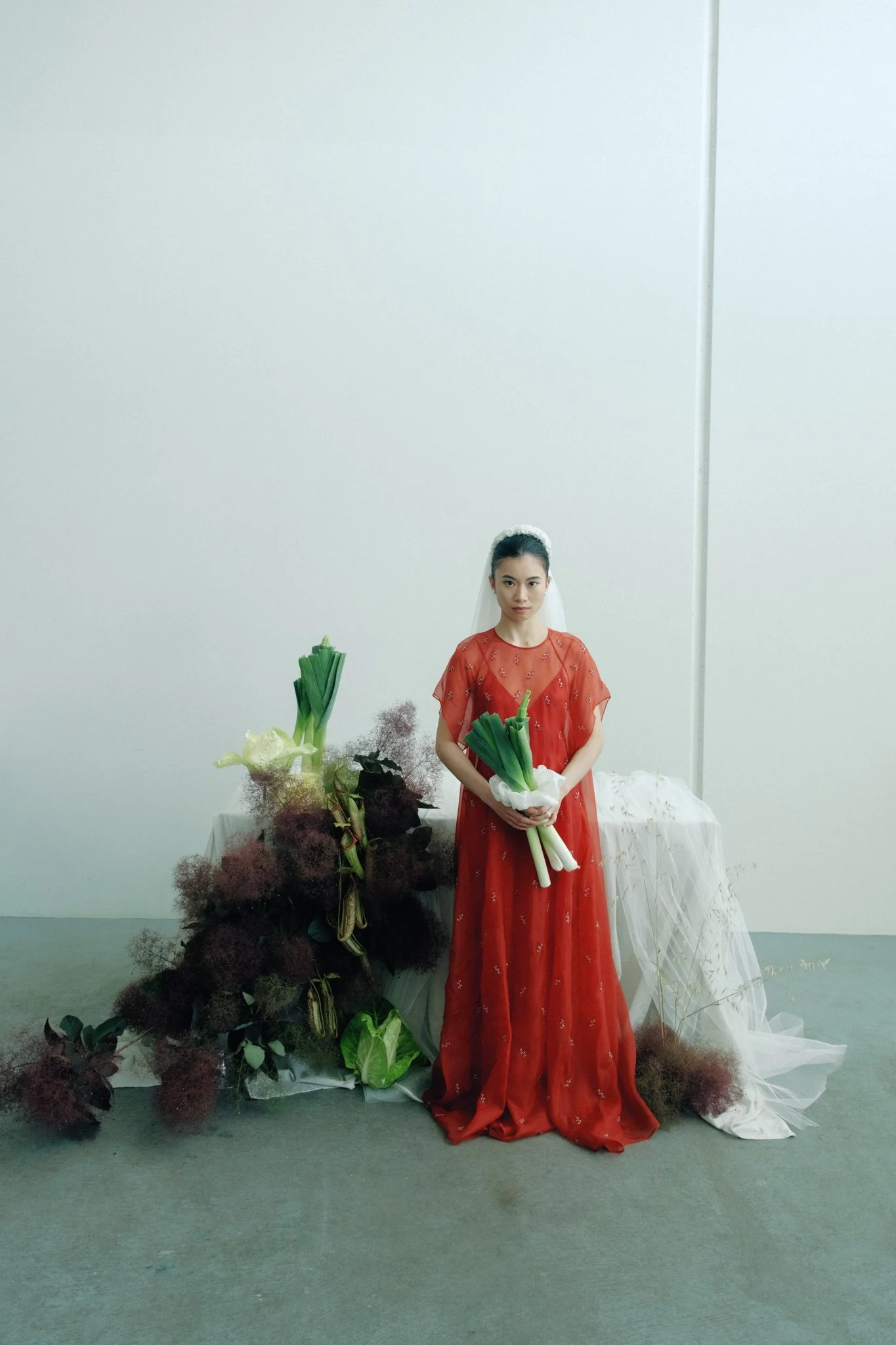 A woman in a red dress standing next to a table filled with flowers and greenery, holding a bouquet of white onions, in a minimalist room with a plain white wall.