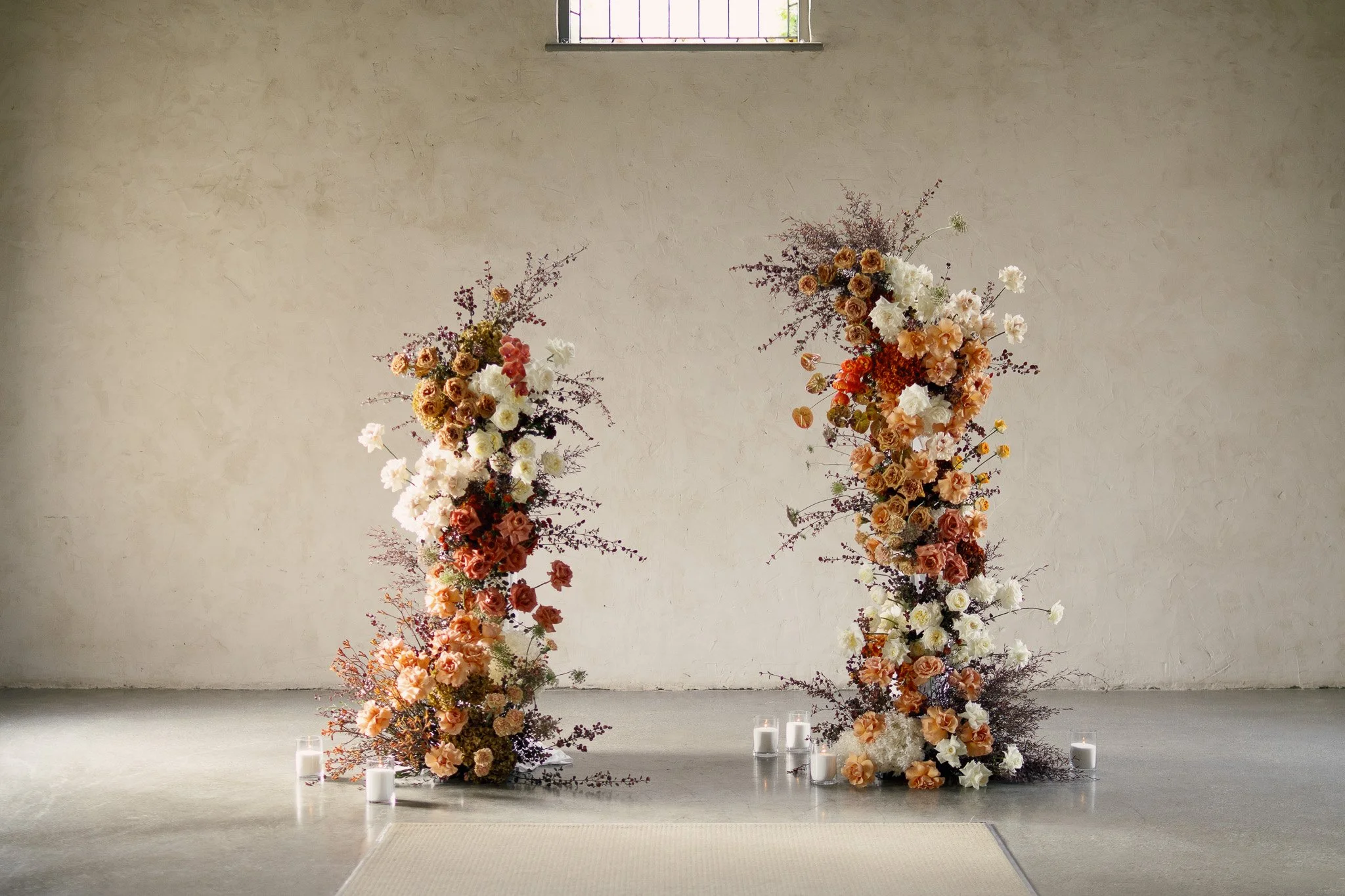 Florist arrangement with roses and flowers in peach, white, and purple tones, with candles at the base, set against a minimalist concrete wall with a small window.