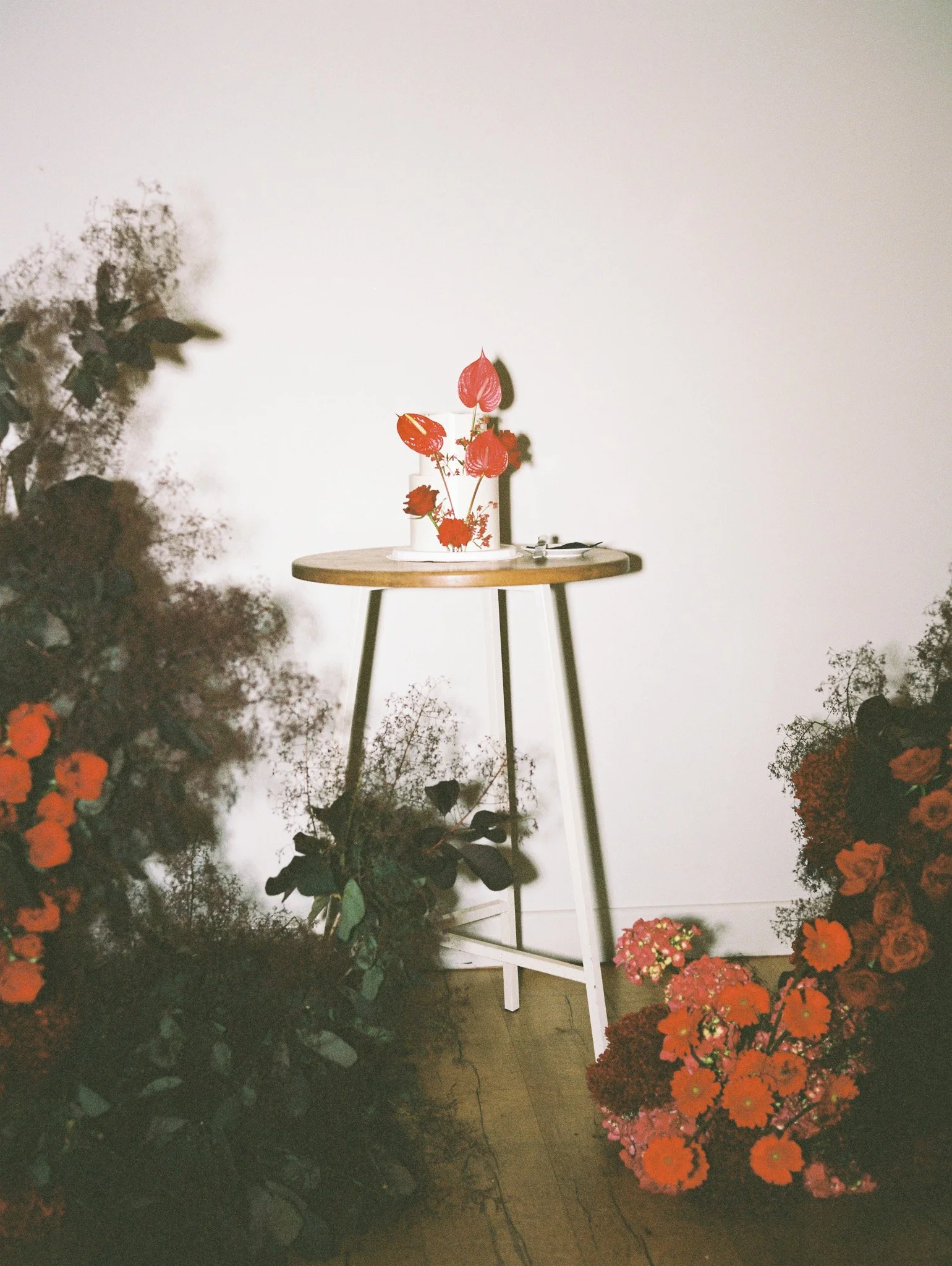 White table with a white vase holding pink and red flowers surrounded by orange and red flowers on the floor, against a plain white wall.
