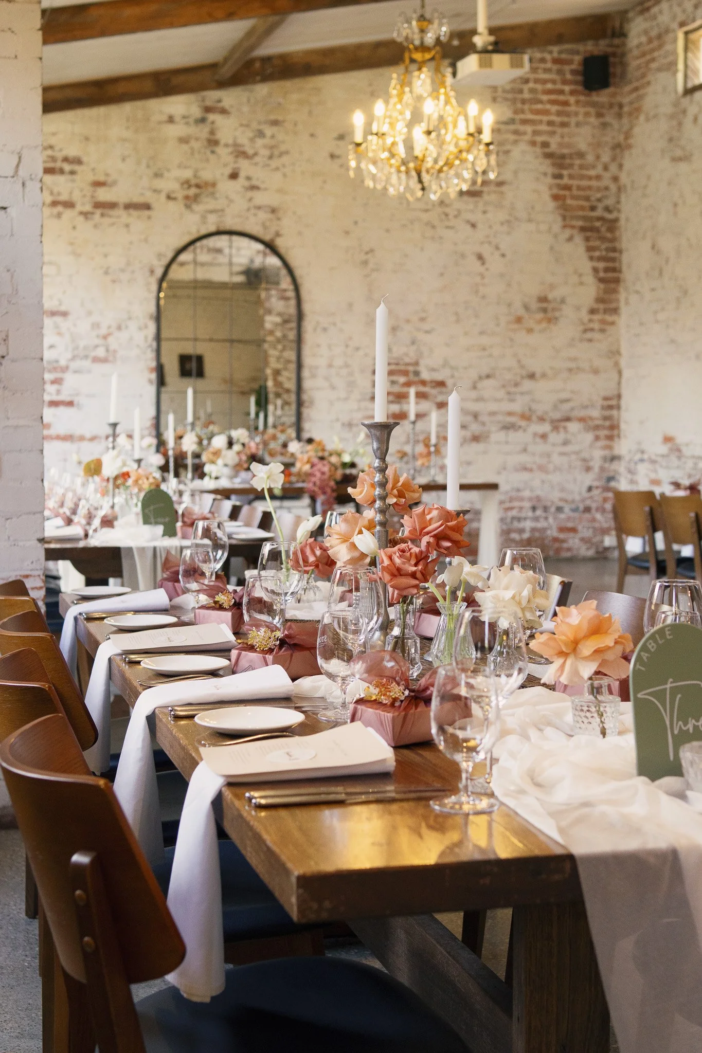 A long wooden table set for a formal event in a rustic brick-walled room. The table has white tablecloths, folded napkins, glassware, and floral centerpieces with peach, white, and pink flowers. Tall white candles in silver holders are also on the ta