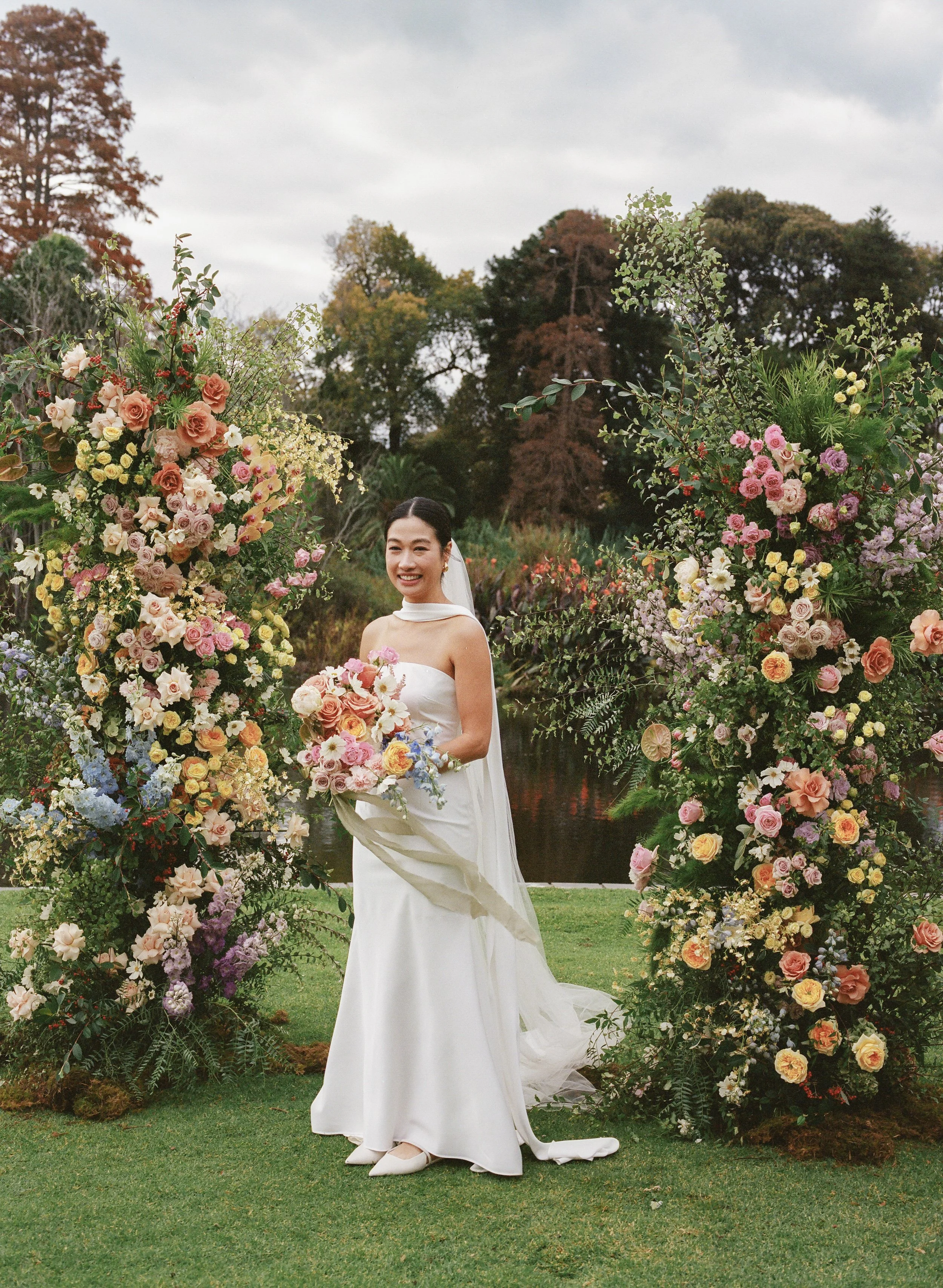 A bride in a white wedding dress standing outdoors. She is smiling and holding a bouquet, surrounded by tall, colorful flower arrangements on either side under an overcast sky.