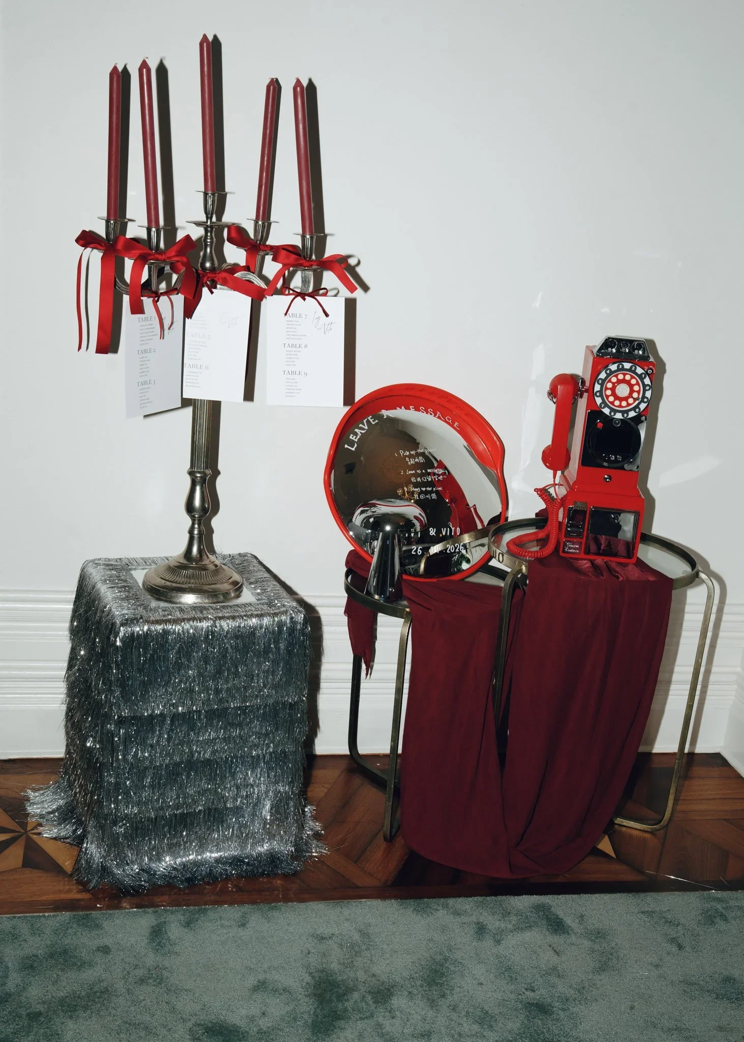 A metallic silver table with a shiny, sparkly silver cloth on top, beside a display of red candlesticks with tags and red ribbons, and a red vintage telephone with a rotary dial and a black and white speech bubble mirror on the floor.
