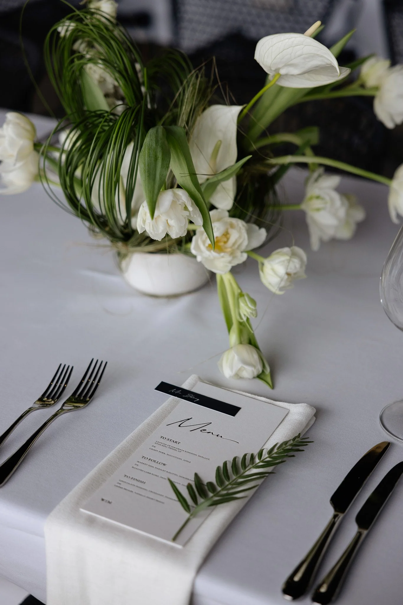 A formal table setting with a white tablecloth, a menu on a napkin, silver forks and knives, and a floral centerpiece with white flowers and greenery.
