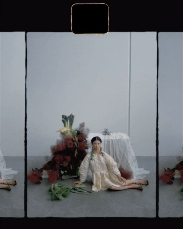 A woman dressed in traditional attire kneeling on the floor in front of floral arrangements and a table with white cloth in a minimalist studio setting.