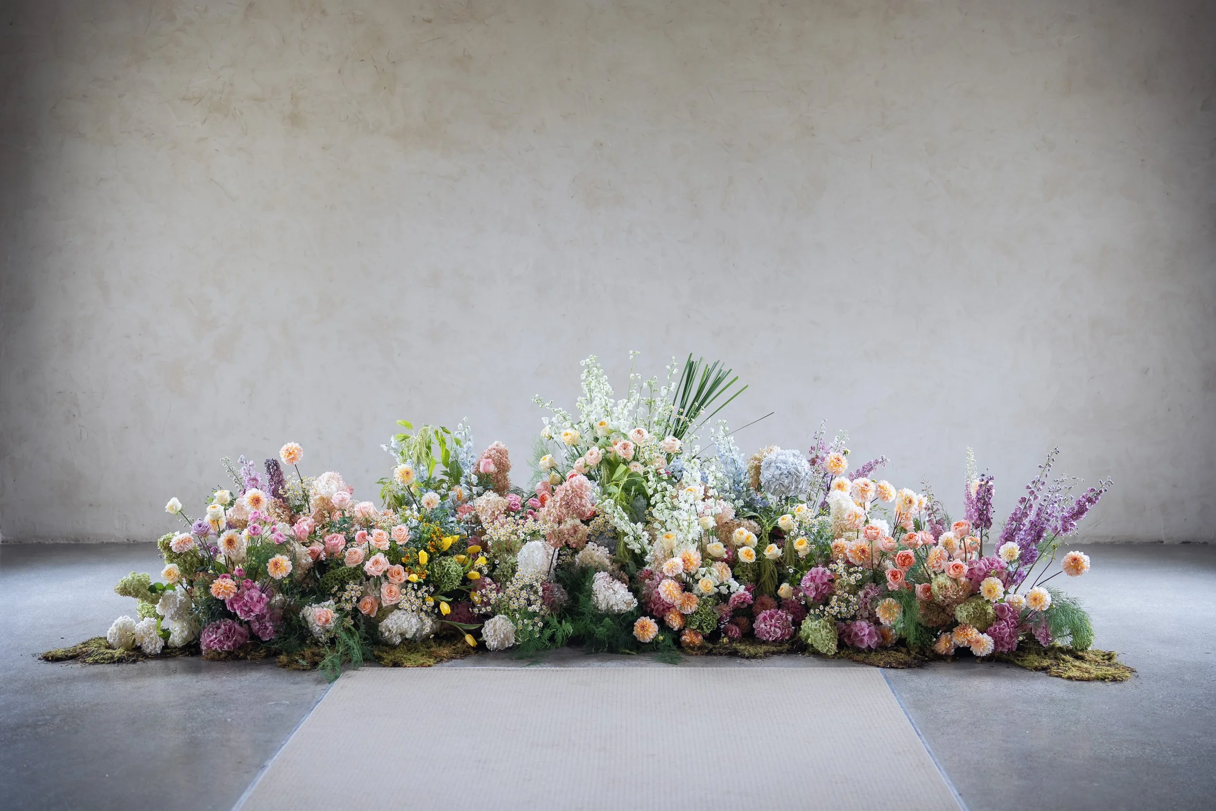 A large arrangement of multicolored flowers including roses, hydrangeas, daisies, and other blossoms, set against a plain wall in an indoor setting.