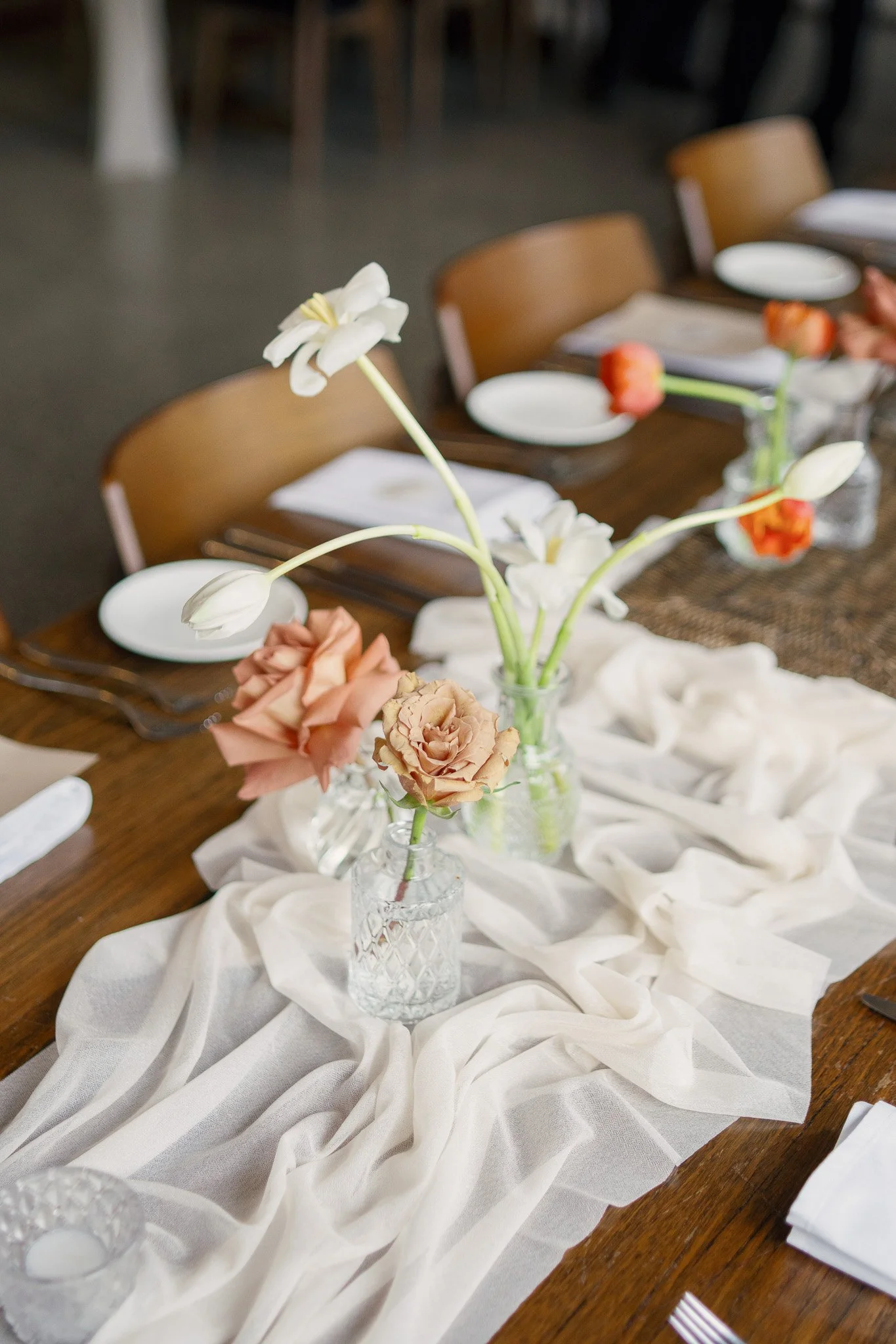 A dining table decorated with vases of flowers, including white lilies and pastel roses, set with white plates, napkins, and silverware on a cream-colored tablecloth.