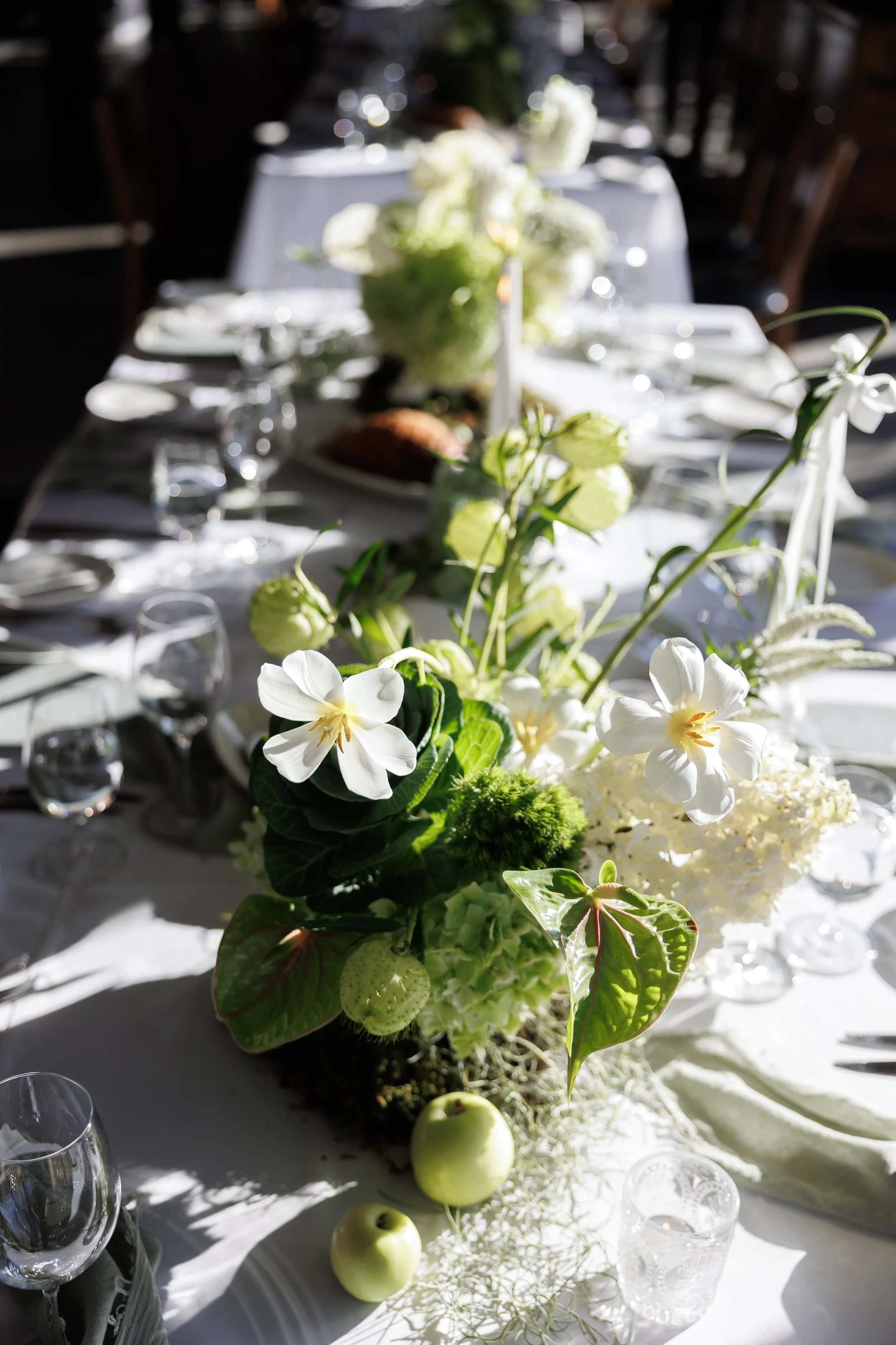 A close-up of a floral centerpiece on a white tablecloth at a dining table, with apples and glassware surrounding it. The flowers are white and green, with some small white blossoms and greenery.