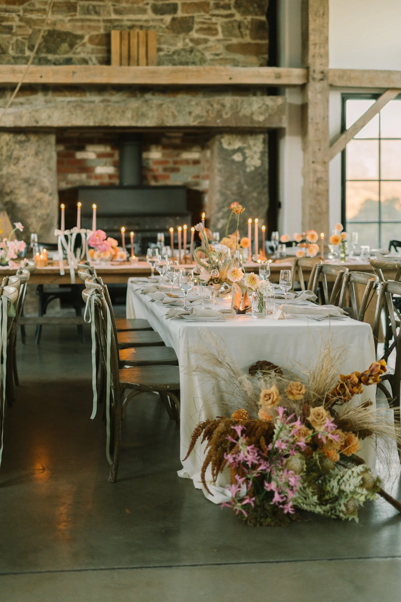 Elegant dining setup with a long table covered in a white tablecloth, adorned with pink and peach flowers, candles, and glassware. The room has rustic stone walls, a fireplace, and large windows with a sunset view.
