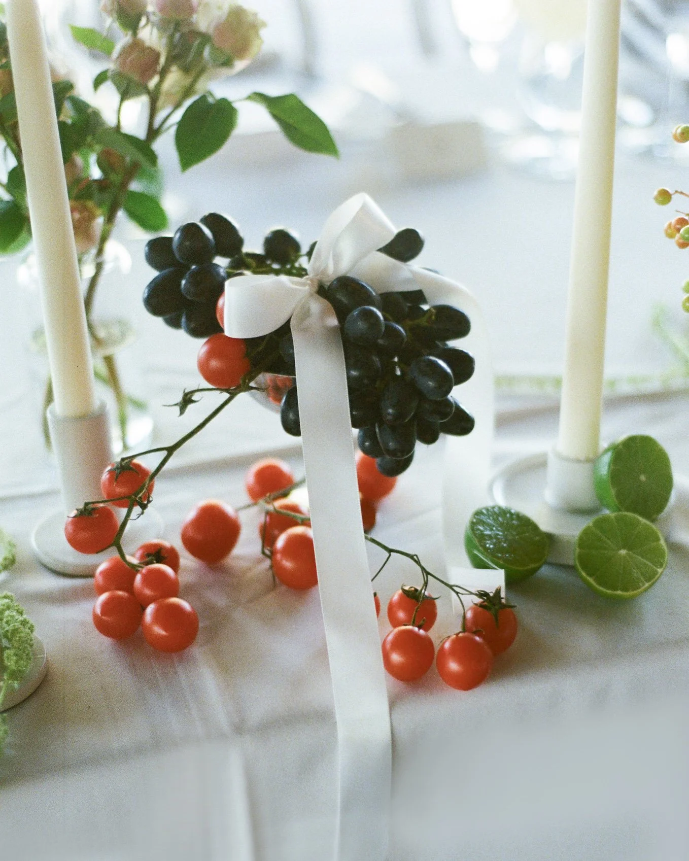 Decorative display of artificial grapes, cherry tomatoes, and green leaves on a white table with two white candle holders.