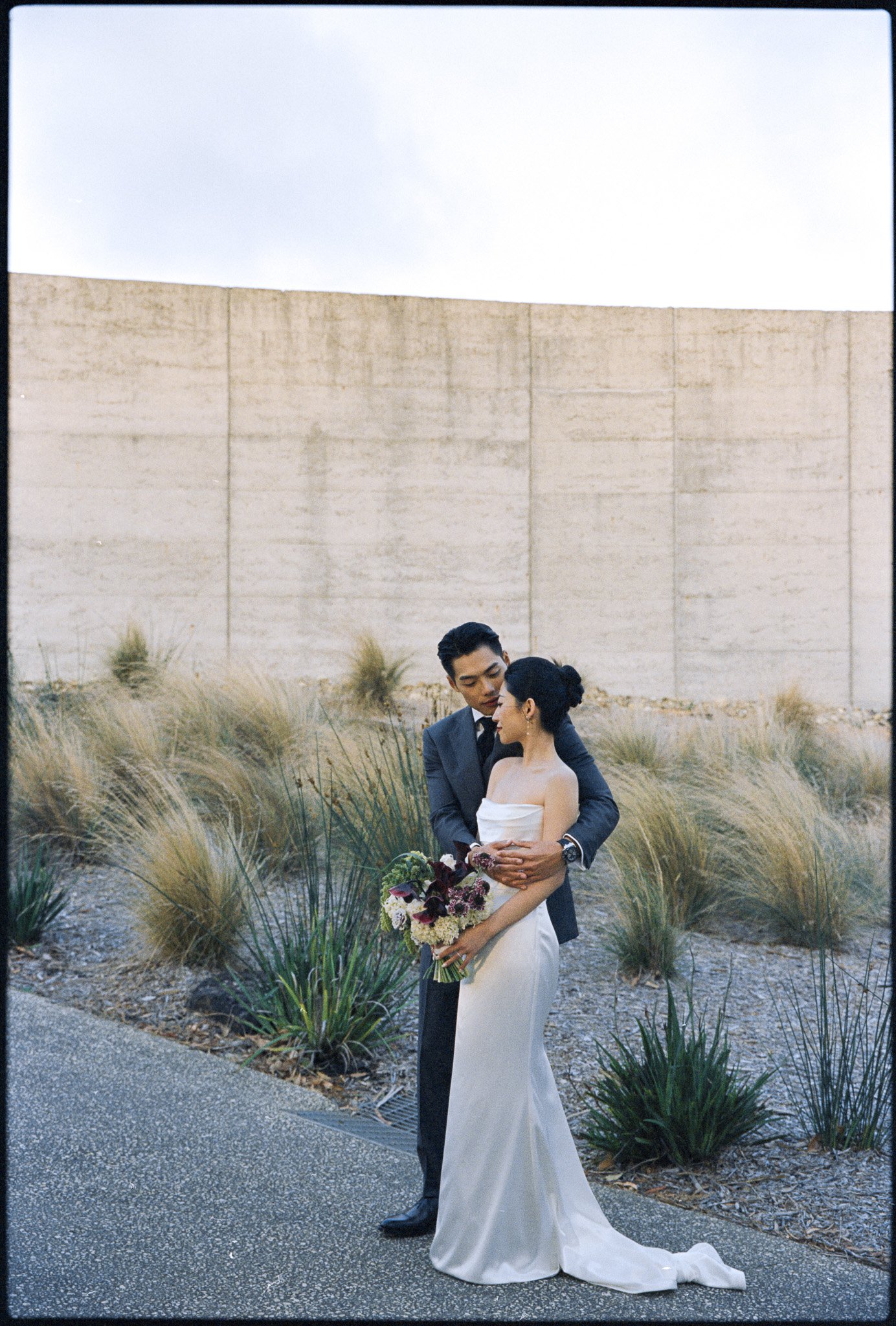 A bride and groom sharing an intimate moment outdoors, surrounded by tall grasses and desert plants, with a beige wall in the background, during a wedding photo shoot.