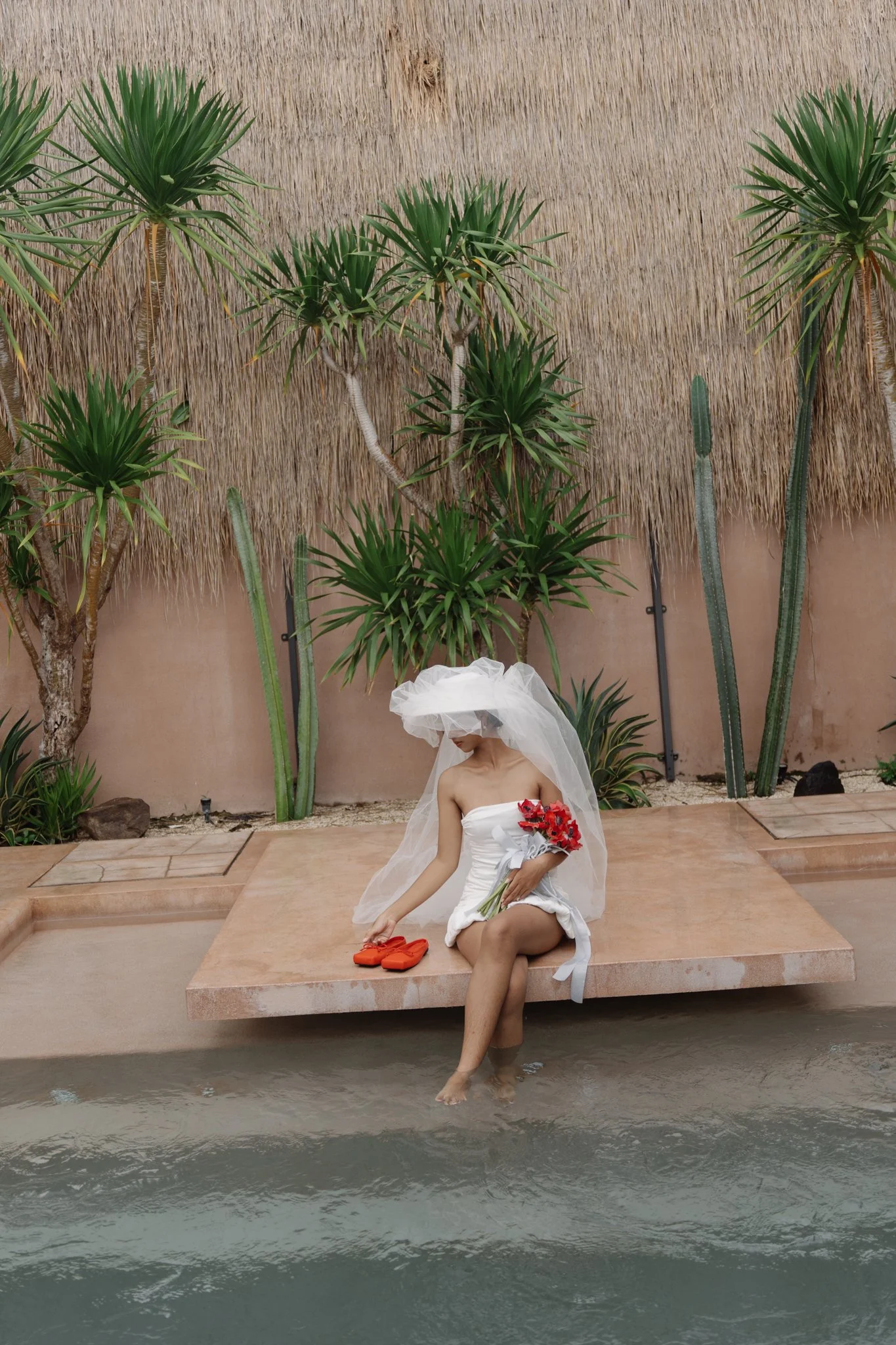 A woman dressed in a white strapless dress and veil sitting on a platform by water, holding red flowers and adjusting orange slippers, surrounded by tropical plants against a thatched wall.