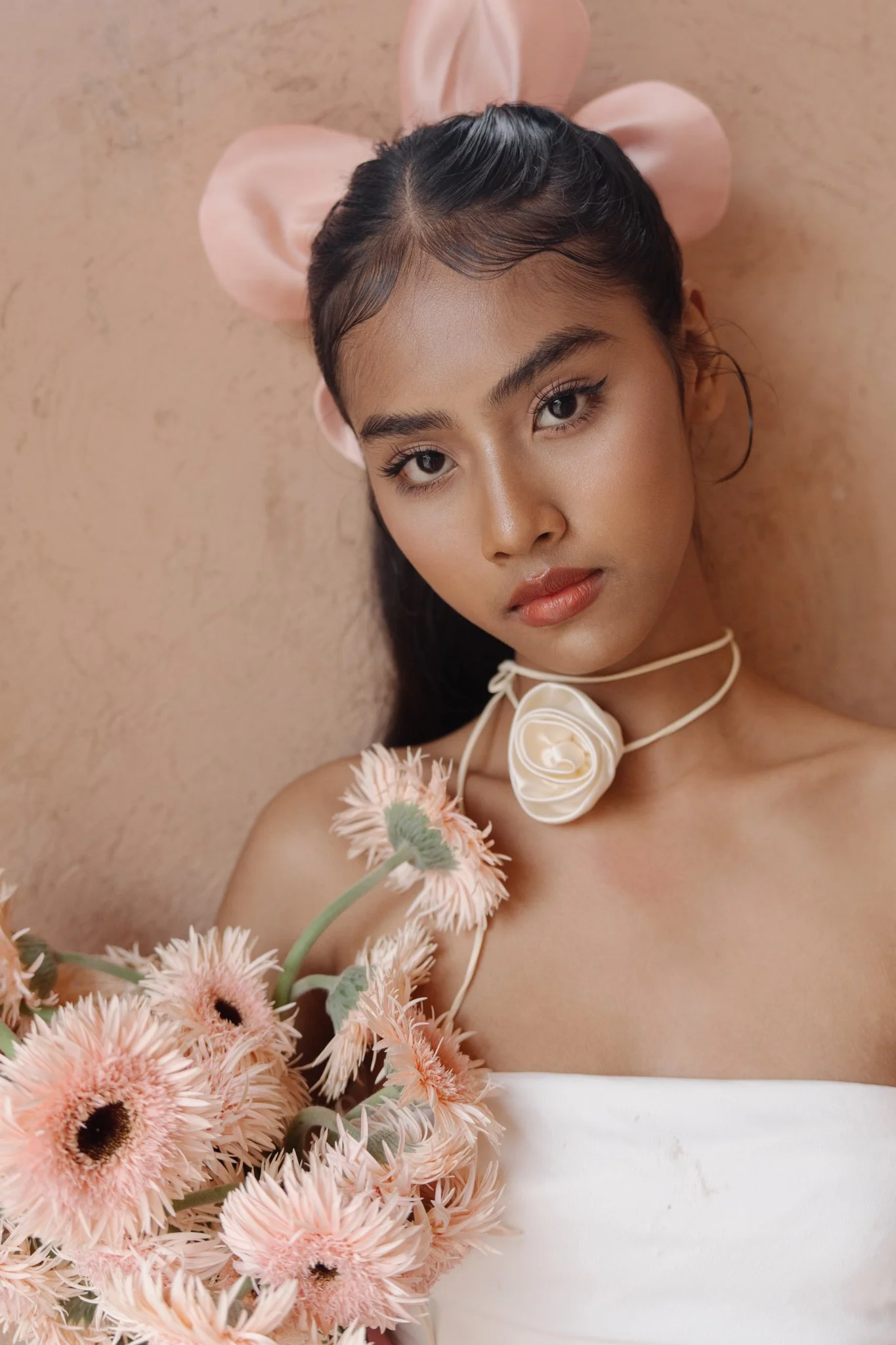 A young woman with dark hair styled in a sleek, wet look, wearing large pink satin bunny ears, a white floral choker, and a white top, lying against a beige wall surrounded by pink and peach flowers.
