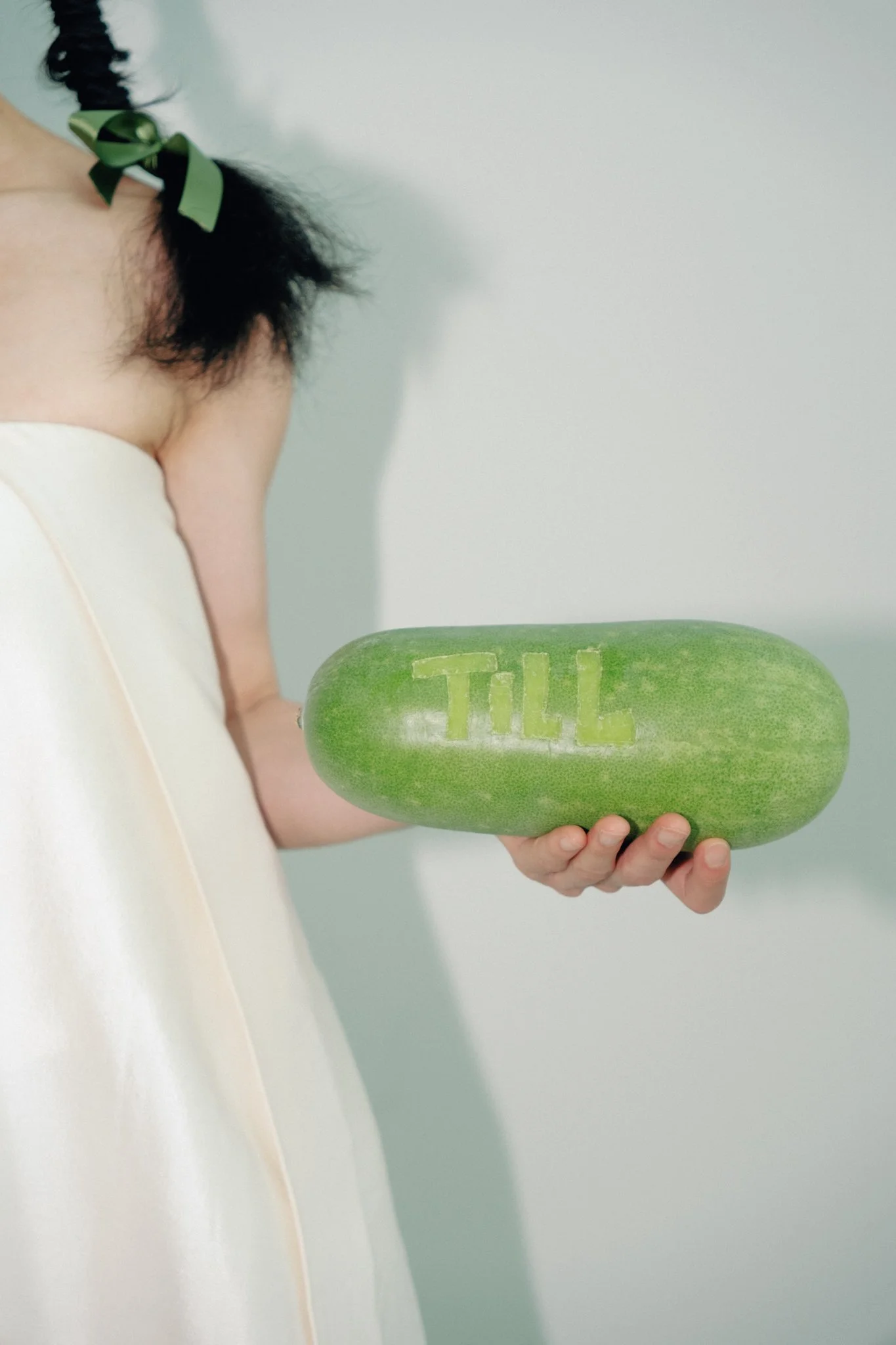 A woman with dark hair in a braid tied with a green ribbon holding a large cucumber with the word 'TILL' carved into it.