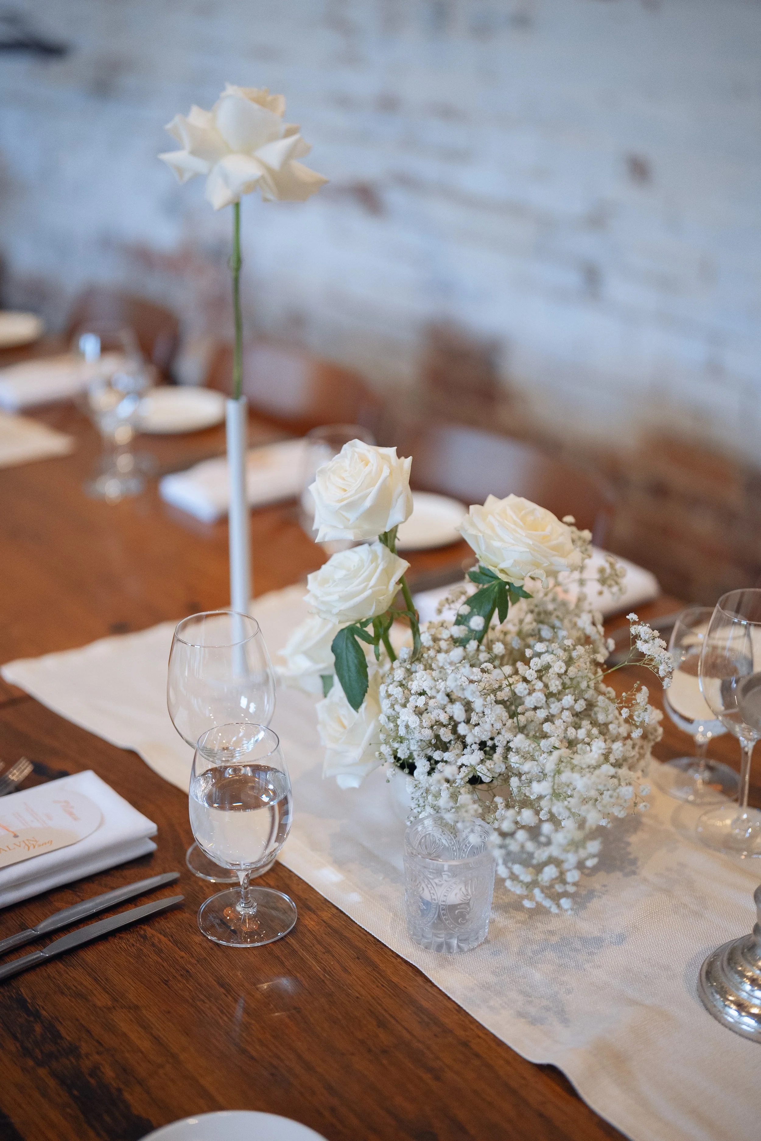 A table set for a formal event with a white floral centerpiece featuring white roses and baby's breath. The table has clear wine glasses, a white linen table runner, and neatly folded white napkins with menus. The background shows a brick wall.