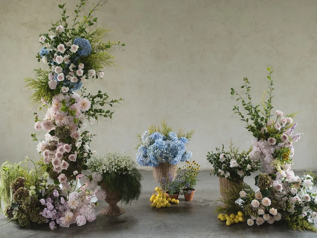 Arrangement of pastel-colored flowers in vases and baskets, with lemons and greenery, in a neutral setting.