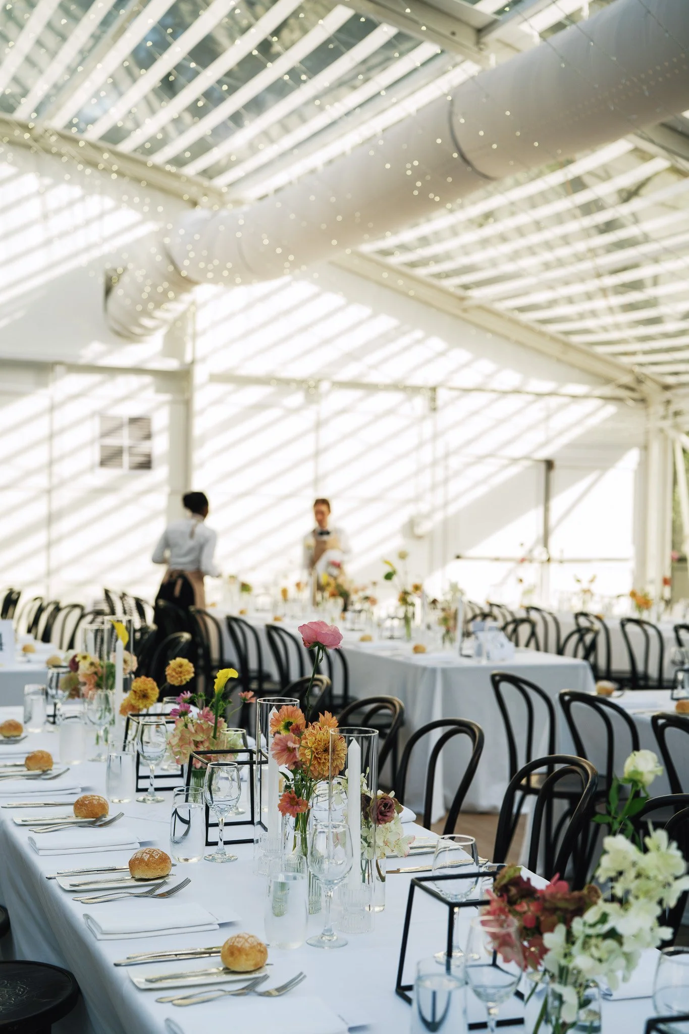 Long banquet table with white tablecloths, decorated with colorful flowers in vases, set with plates, glasses, utensils, and bread rolls. The setting appears to be in a bright, airy venue with a high ceiling and glass roof, letting in natural light, 