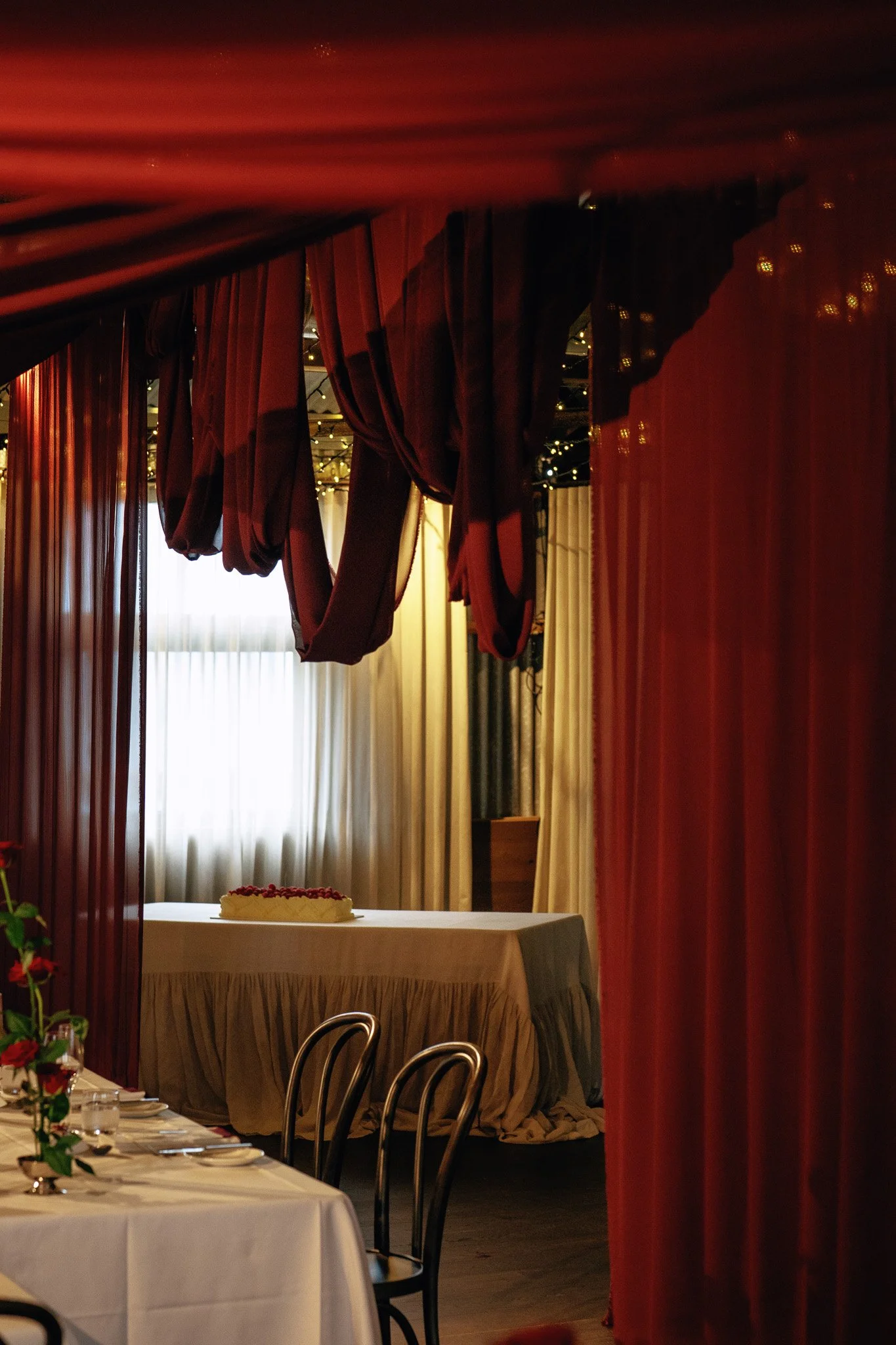 Indoor party or event space with decorated table, cake, and elegant curtains with string lights.