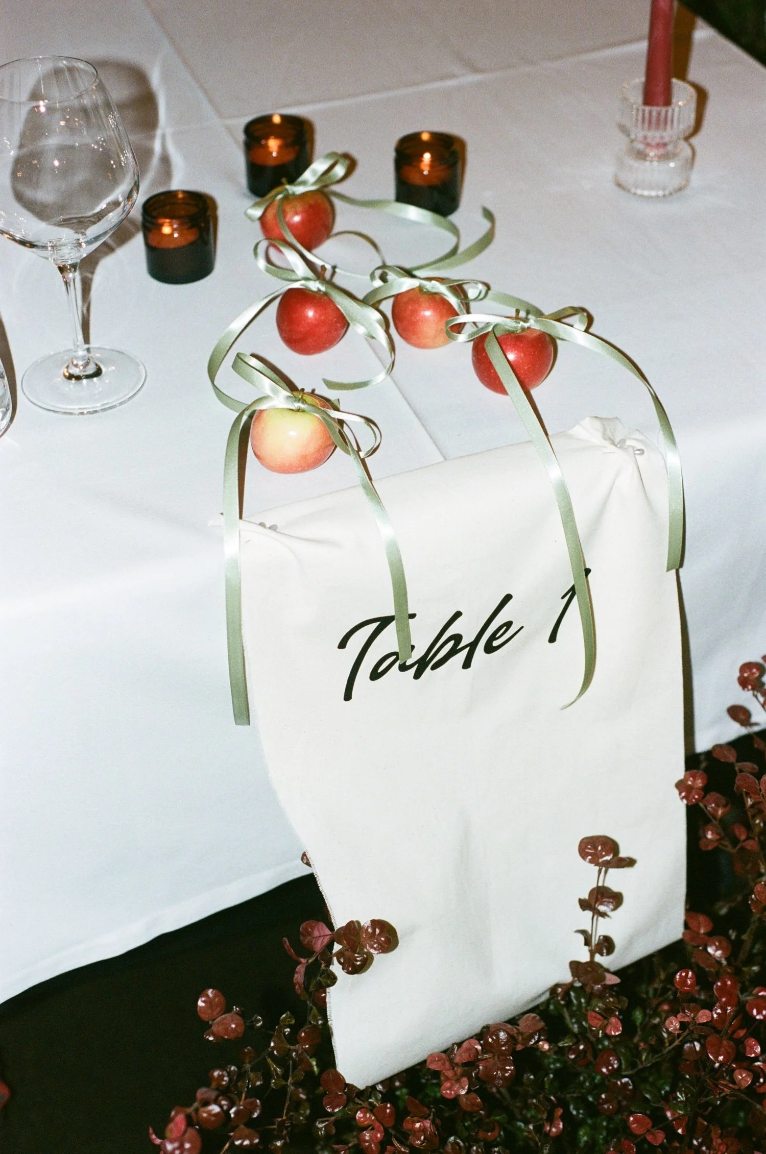 Table decoration with apples, green ribbons, chocolate votive candles, and a candle in a glass holder. White tablecloth with a sign that partially reads "Table".