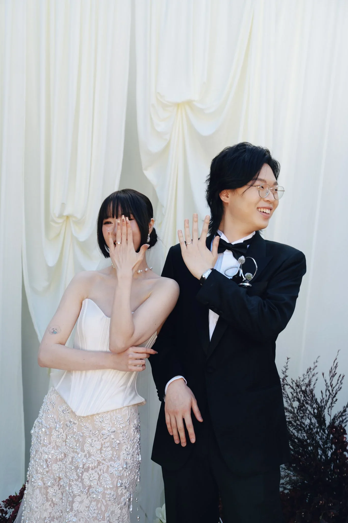 A happy bride and groom standing in front of cream-colored drapes, smiling, with the bride covering her face with her hand and the groom showing his ring finger.