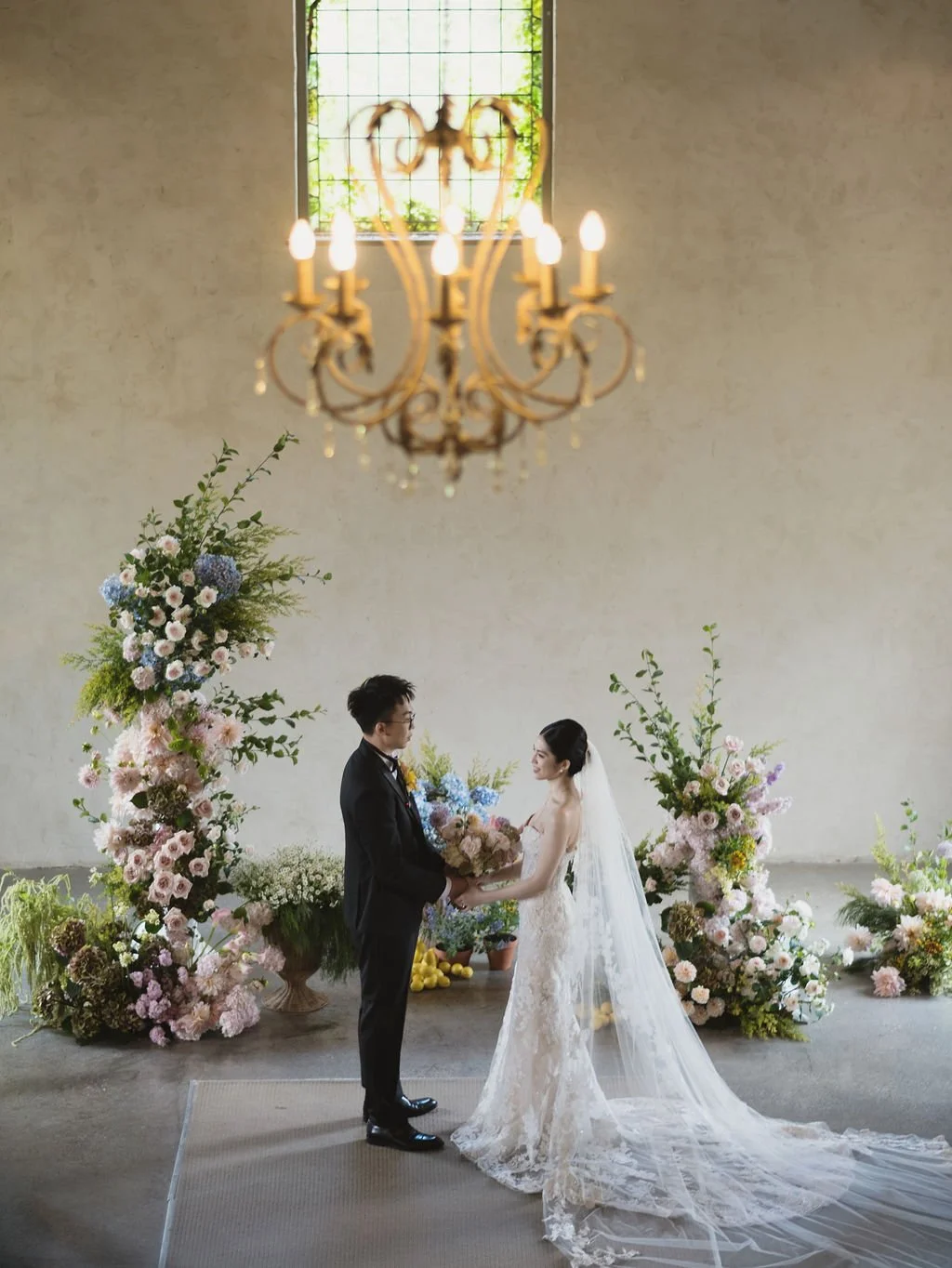 A bride and groom standing in front of a floral arch, exchanging a bouquet at their wedding ceremony inside a church with a chandelier hanging above.