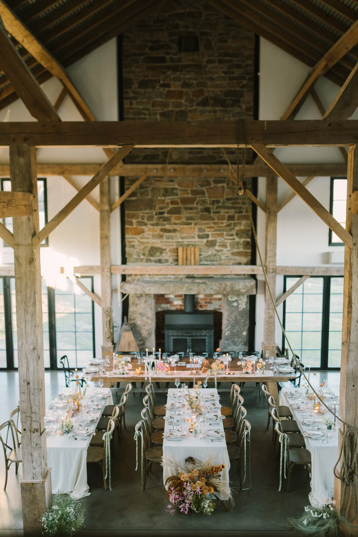 Interior of a rustic wedding venue with long dining tables decorated with flowers, candles, and table settings, set beneath a high wooden ceiling with exposed beams and a stone fireplace at the end.
