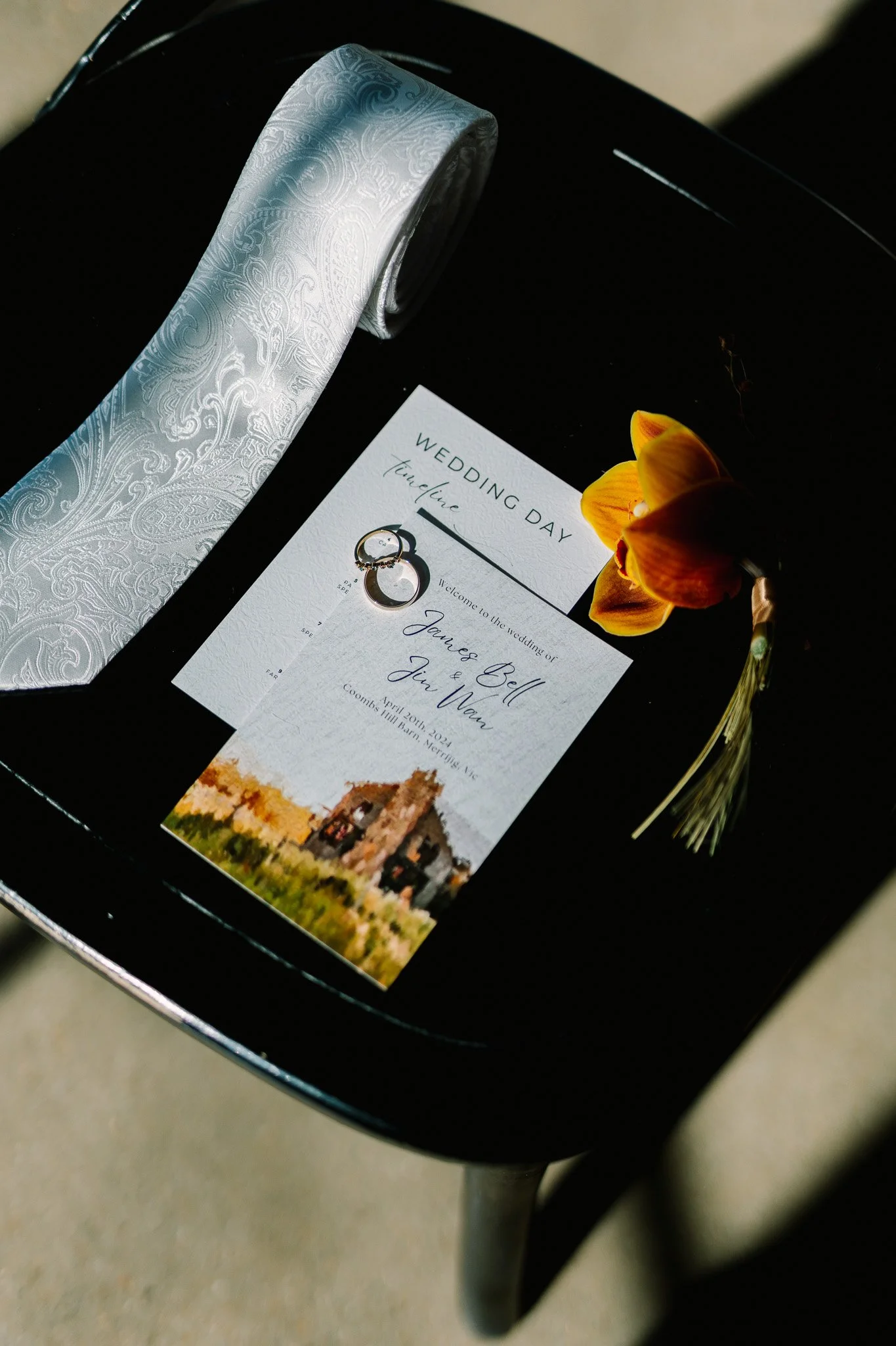 Wedding invitation, two wedding rings, yellow flower, and a rolled silk ribbon on a black surface.
