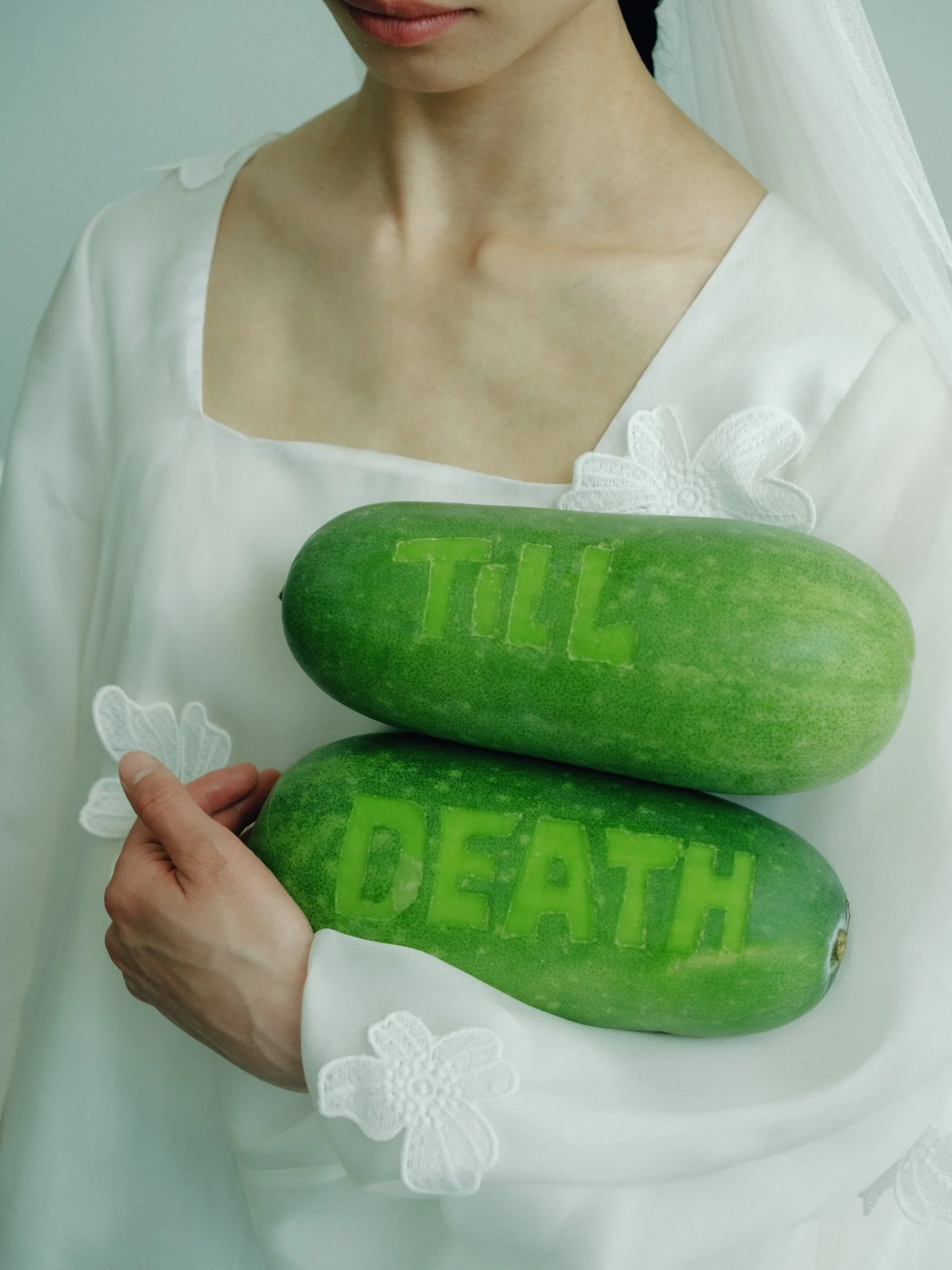 Close-up of a woman wearing a white dress with floral embroidery holding three stacked green rocks with the word 'DEATH' carved into the top one. The top of her face is partially visible, showing her lips and chin.