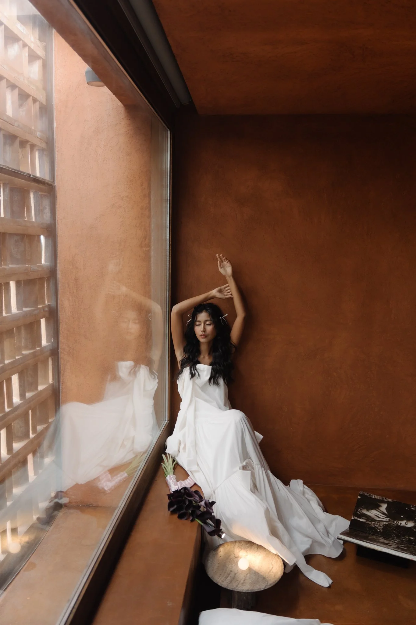 A woman dressed in a white gown sitting on the floor of a corner, with her eyes closed and arms raised above her head, next to a window reflecting her image, with a bouquet of purple flowers on the floor and a black and white book beside her.